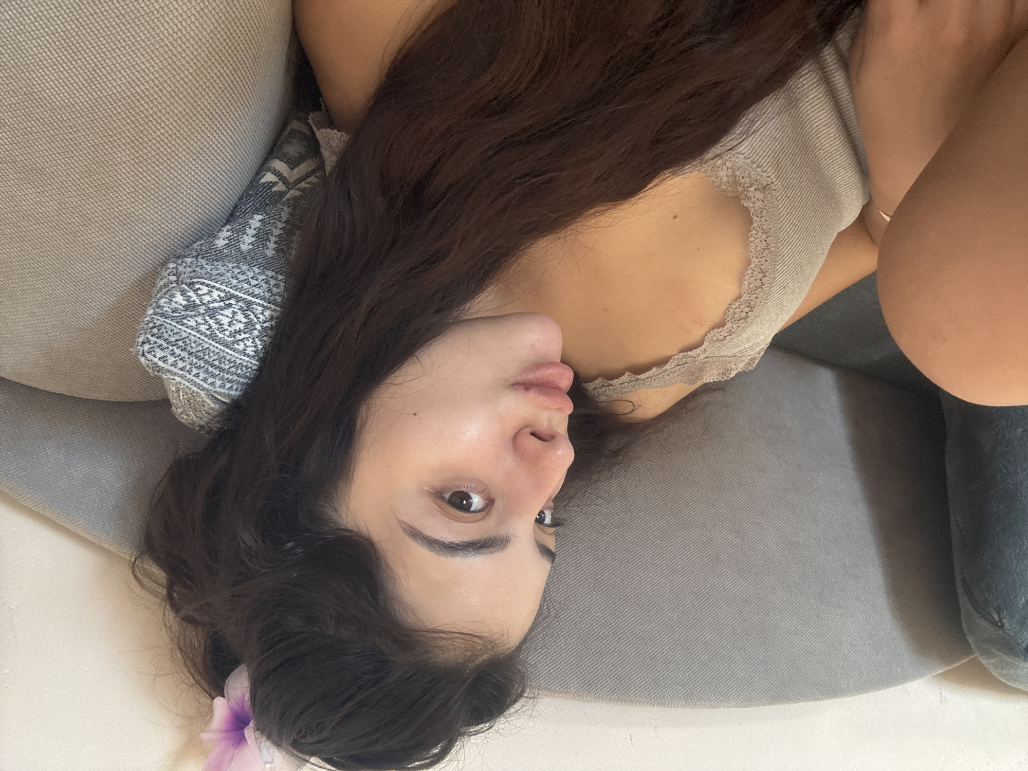 A woman lies on a gray sofa, her head resting on a patterned pillow. She is wearing a beige top with lace trim. Her dark hair is loose, with a purple flower in it. The background is neutral. This scene does not appear to be related to Plovdiv, Bulgaria.