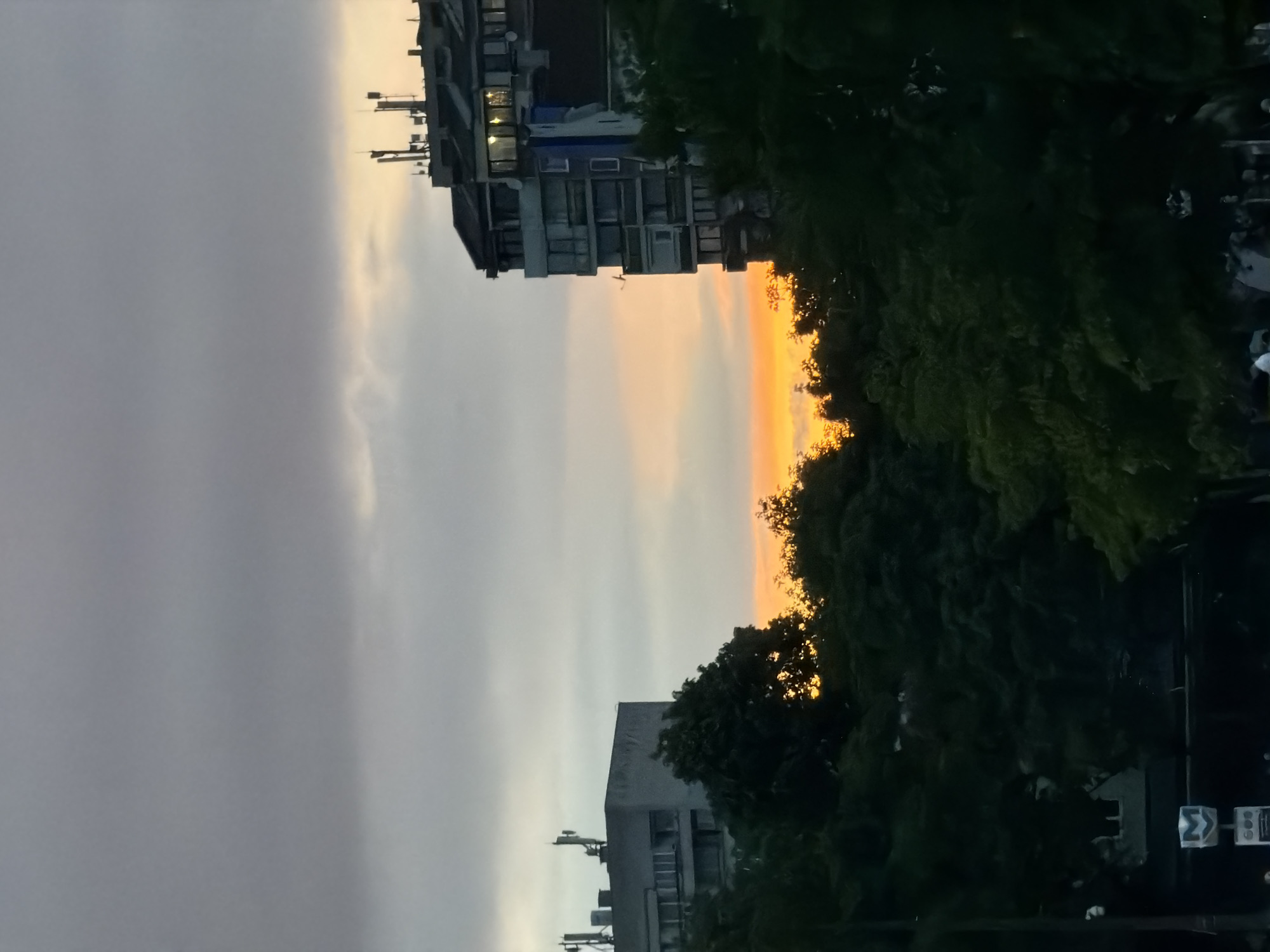 The image captures a cityscape at dusk in Sofia, Bulgaria. Two multistory buildings are visible on either side of a narrow sky view. The sky displays a gradient of colors from grey clouds on the left to orange near the horizon. Trees with dark foliage frame the skyline on the right. Communication towers are seen on the top of the buildings. At the bottom right, there is a road sign with a blue "sigma" symbol, indicating potential traffic or directional information.
