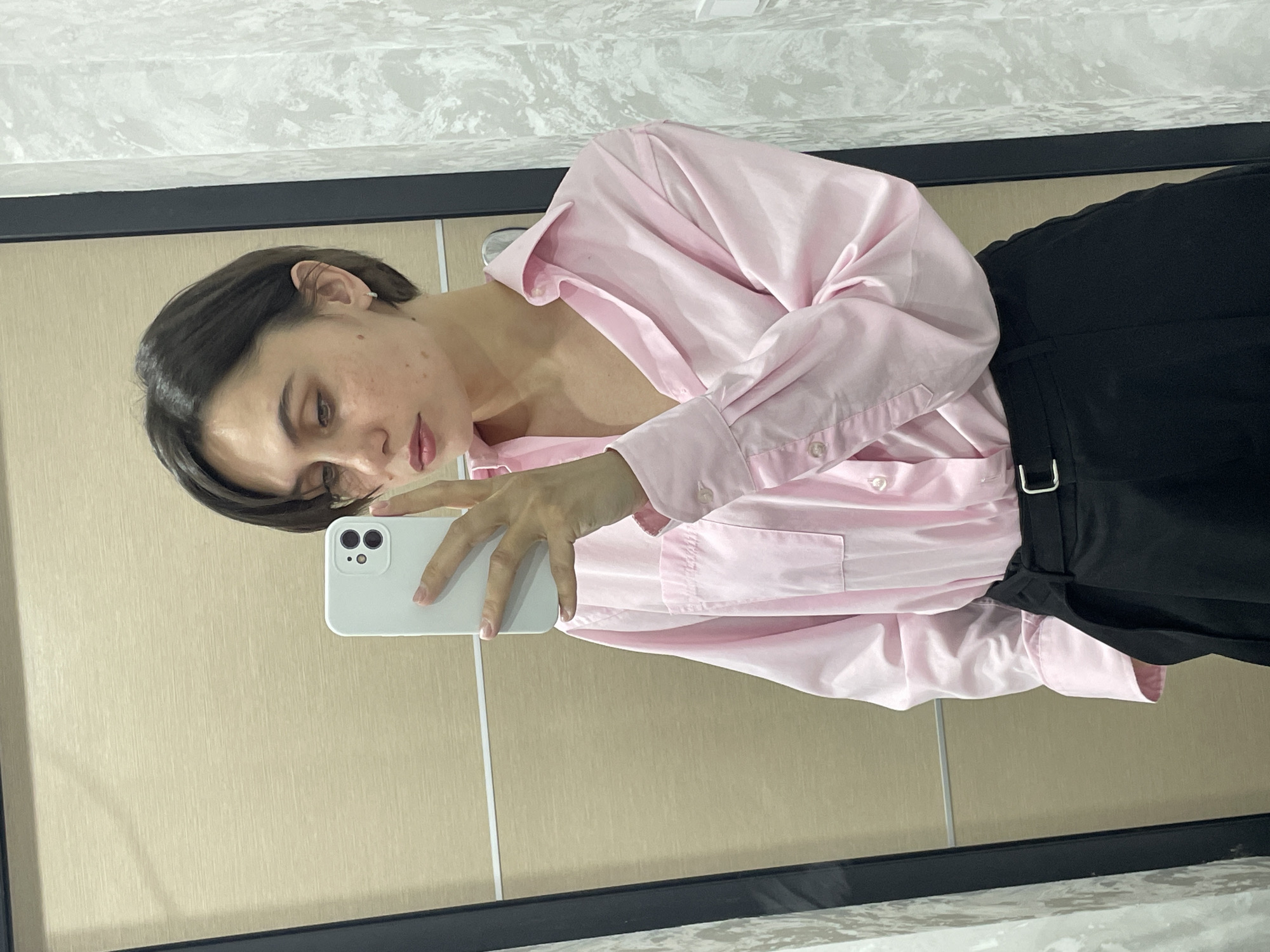 A woman with short brown hair is taking a selfie in a mirror. She is wearing a pink button-down shirt and black pants. The background appears to be a wall and a countertop. There is no indication of this being located in Izhevsk, Russia.