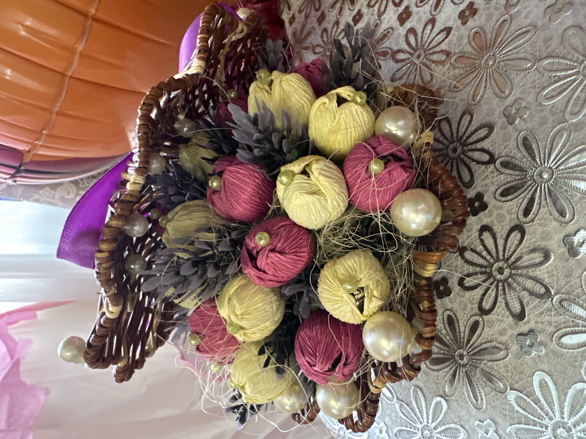 The image captures a close-up view of a decorative arrangement, likely placed indoors. The central subject is a woven basket, crafted from brown material, containing an assortment of artificial elements.
Within the basket, there are numerous fabric "flowers" or buds, predominantly in shades of yellow and magenta/maroon. Each of these fabric elements is adorned with a small, light green bead at its center, resembling a pistil. Interspersed among these fabric blooms are dried or artificial plant elements in a grayish-purple hue, along with strands of light-colored, thin fibers or raffia. Several large, lustrous, pearl-like beads are also integrated into the arrangement, enhancing its decorative quality. A purple ribbon is tied to the basket, adding a pop of color and a festive touch.
In the background, a light-colored surface with a distinct floral pattern is visible, resembling a tablecloth or decorative fabric. The pattern consists of repeating flower motifs in shades of white, silver, and light brown. To the upper left, an orange, ridged object, possibly an inflatable item like a balloon or part of a piece of furniture, is partially visible. The lower left corner shows a soft pink fabric.
No people or activities are present in the image, focusing solely on the decorative object. The setting appears to be a well-lit indoor environment. Partial, indistinct text is visible on one of the yellow fabric flowers, though it is largely obscured and unreadable. The arrangement suggests a celebratory or decorative context, possibly for an event or home decor in Yaroslavka, Russia.