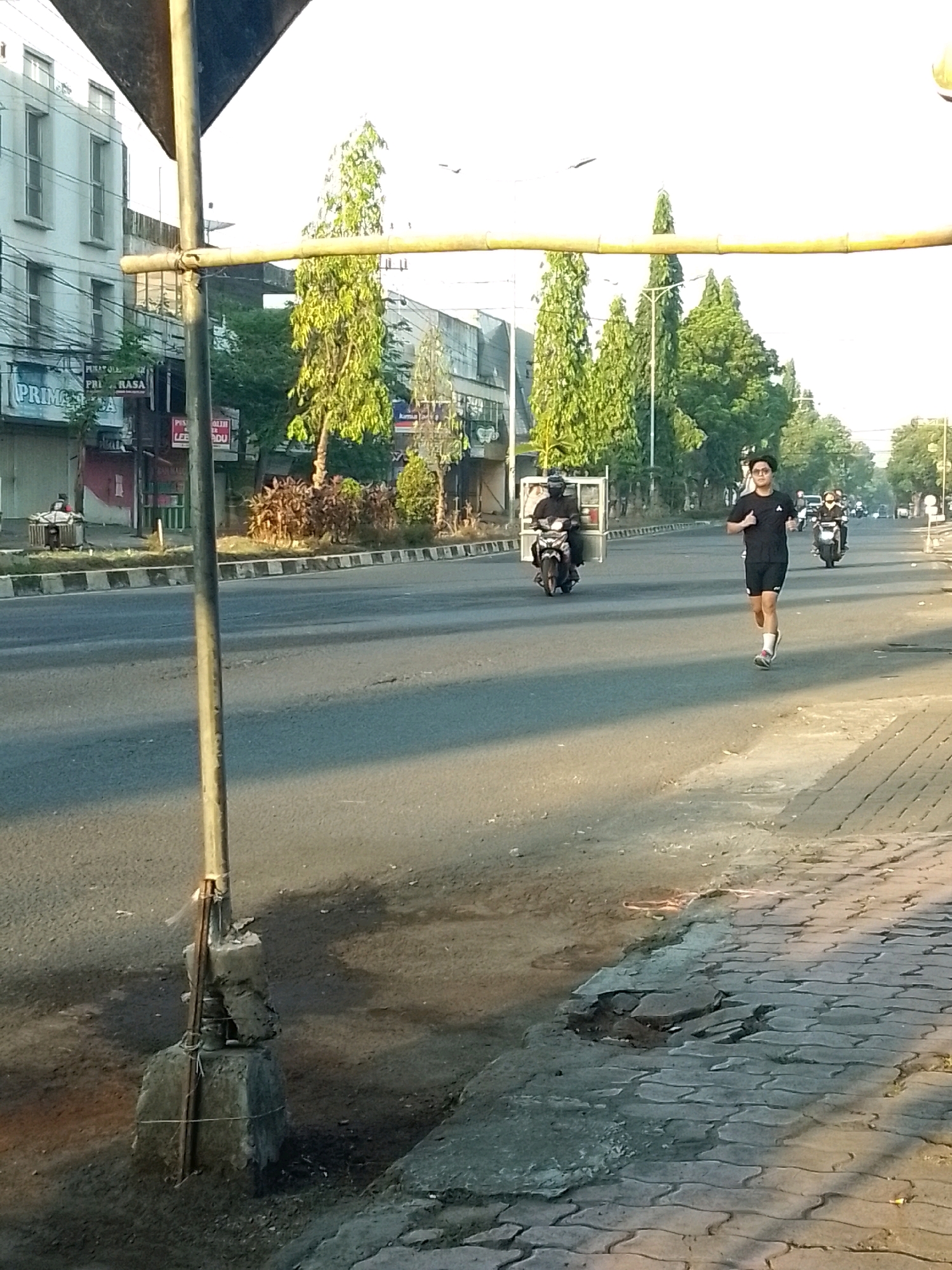 The image depicts a street scene in Jember, Indonesia, during daylight. A young man is jogging on the right side of the road, wearing a black t-shirt and shorts. He is running on a paved sidewalk. In the background, several motorcycles are visible, some with riders. One motorcycle is on the road to the left of the jogger, and another is further down the road. Buildings line the left side of the street, including a white building with multiple windows. Trees and foliage are present along the street and in front of the buildings. A metal pole with a bamboo crossbar stands in the foreground on the left, attached to a concrete base. The road surface appears to be asphalt with some cracks and discoloration. The sidewalk on the right is paved with interlocking bricks, and parts of it are broken or damaged. The sky is bright and clear, suggesting it is a sunny day.