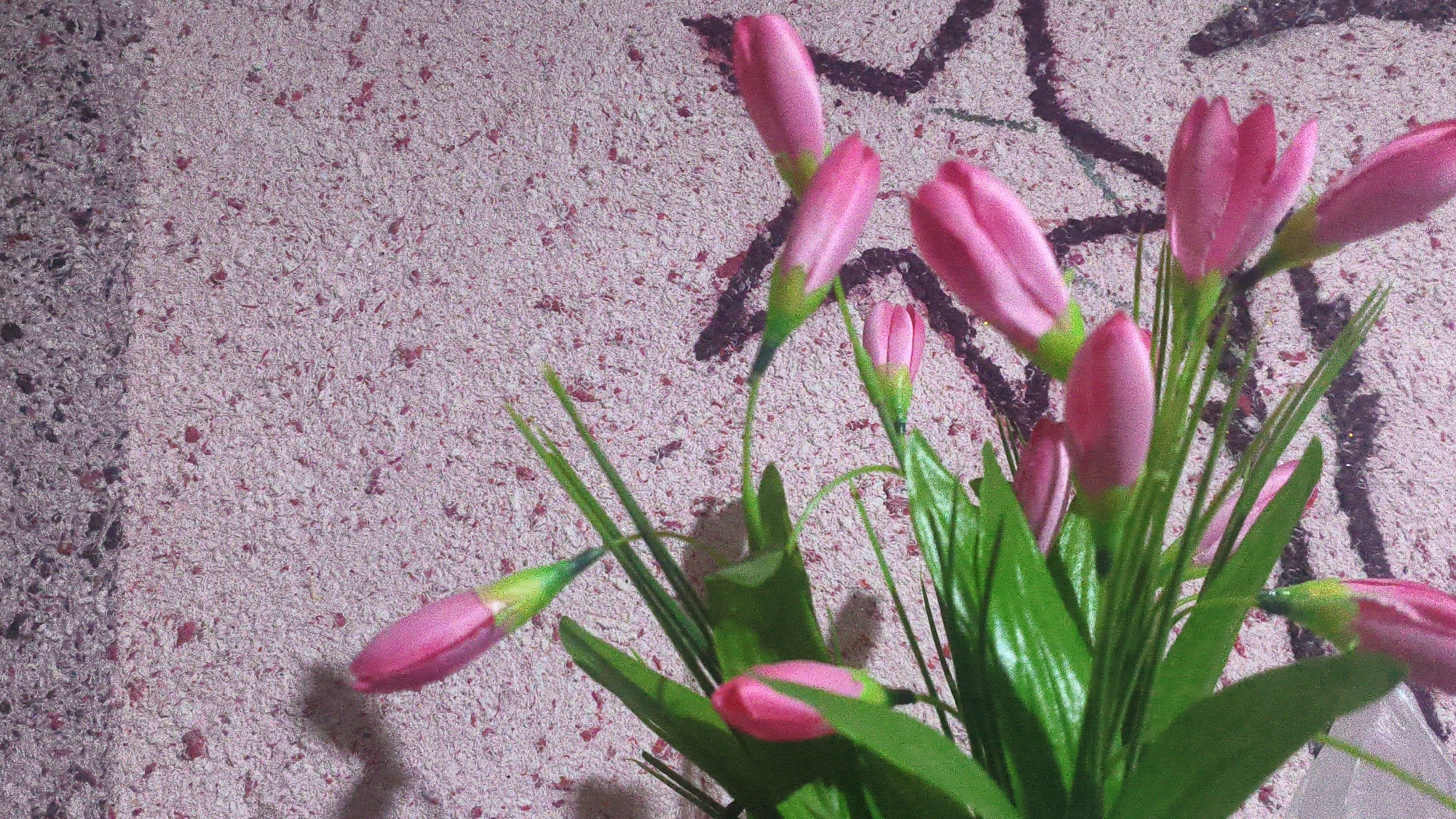 The image shows a close-up view of artificial pink flowers with green stems and leaves against a textured pink and purple background. The background appears to be a wall with a rough finish and some dark, abstract markings. The lighting suggests an indoor setting, with a shadow cast by the flowers on the left side of the image. There are no people visible in the scene, and the focus is entirely on the floral arrangement. The image quality is somewhat grainy, possibly due to low light or the texture of the surface. The scene does not depict any specific urban environment, including Mehnatabad, Uzbekistan.