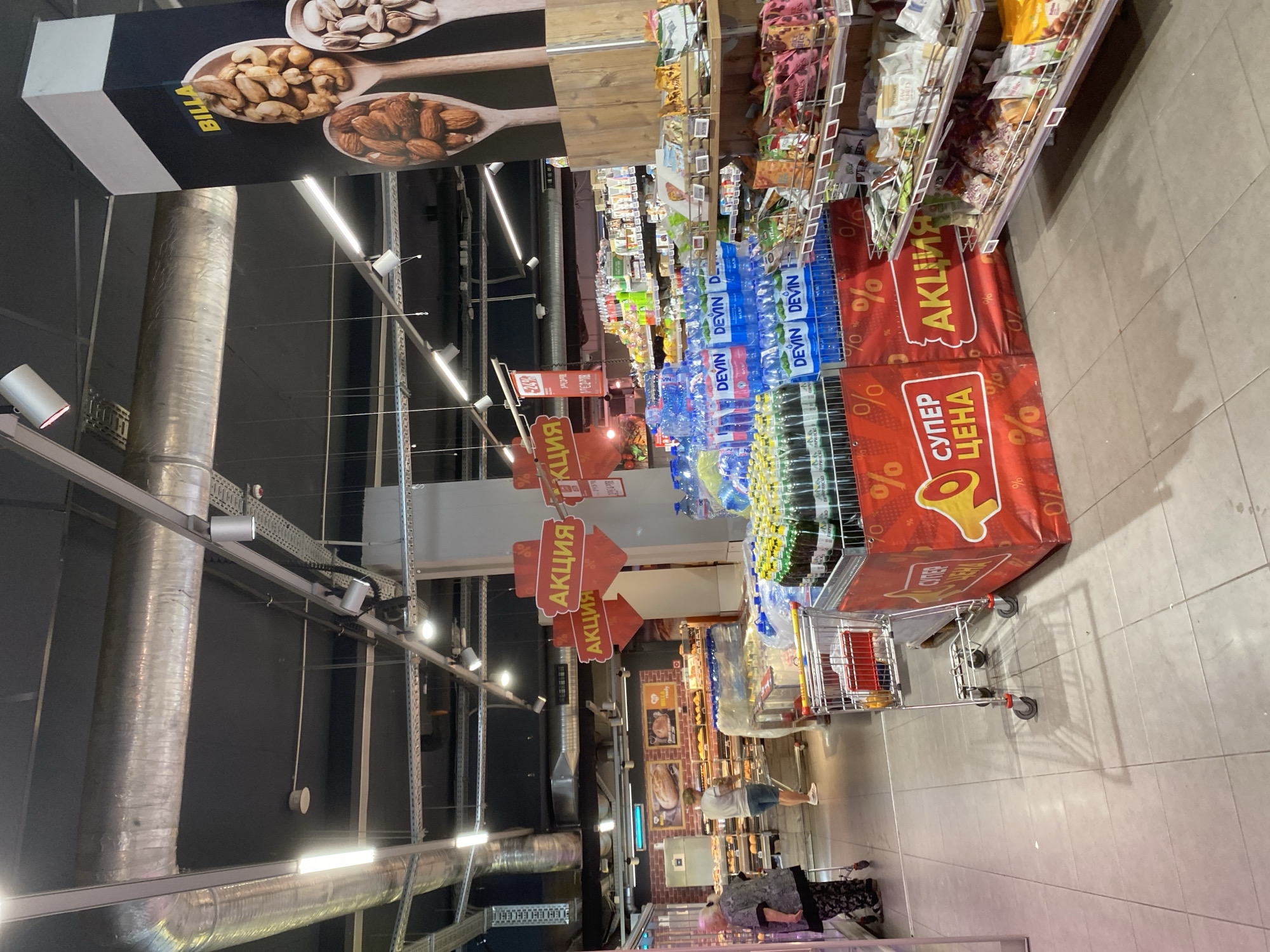 The image depicts the interior of a Billa supermarket in Varna, Bulgaria. In the foreground, a shopping cart is partially visible. To the right, shelves are stocked with various packaged goods, including snacks and nuts. A prominent red banner with "АКЦИЯ" (Action/Sale) and "СУПЕР ЦЕНА" (Super Price) text, along with percentage symbols, indicates a promotional event. Bottled water, labeled "DEVIN," is stacked in the middle ground. In the background, customers are browsing in the bakery section, which features shelves of bread and pastries. Overhead, industrial lighting and ventilation systems are visible. A large banner with images of nuts and the "BILLA" logo is positioned on the right side of the frame.