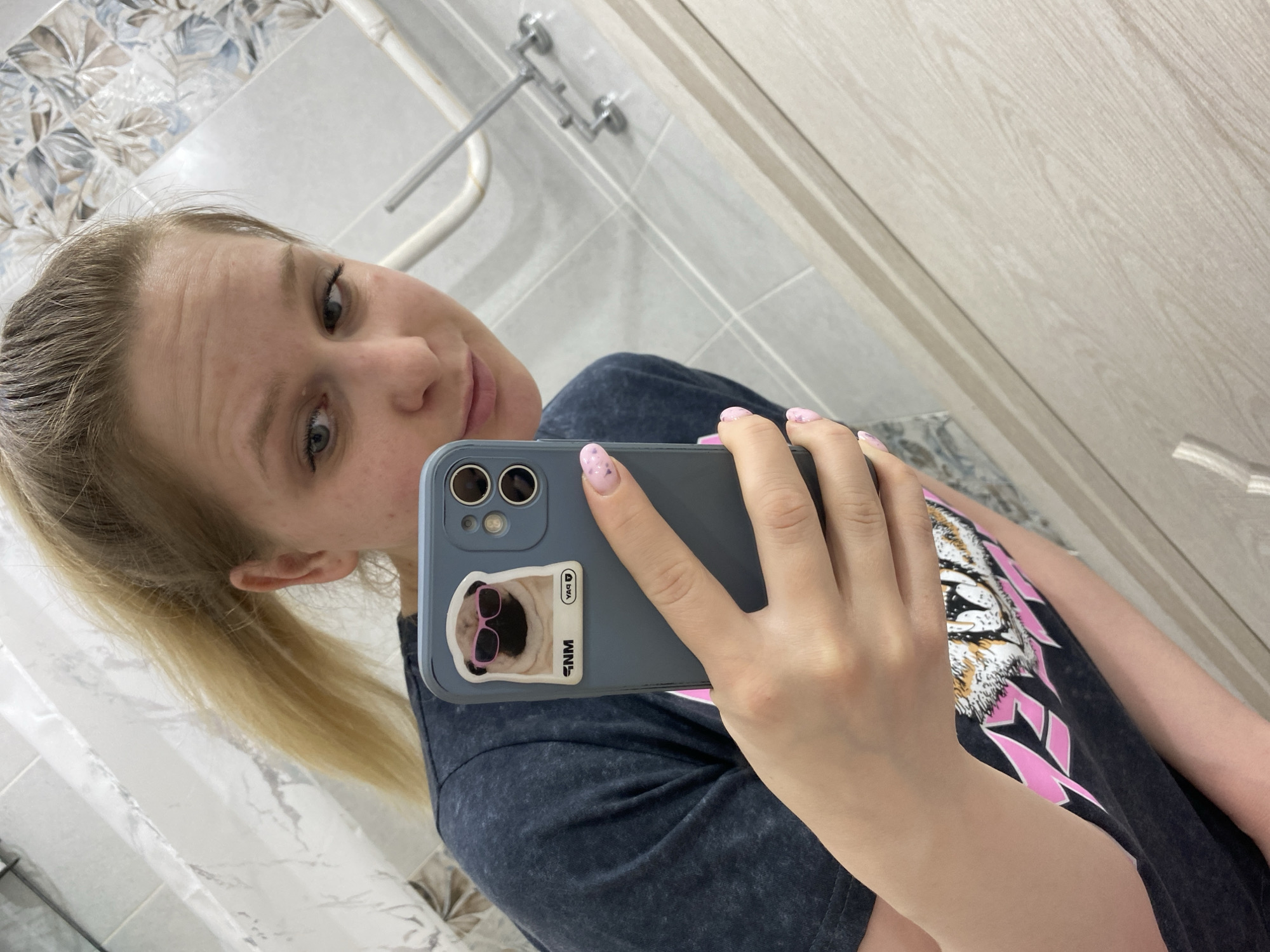 The image shows a woman taking a selfie in a bathroom. The bathroom features a showerhead and patterned wall tiles. She has blonde hair pulled back in a ponytail and is wearing a dark t-shirt with a tiger graphic and pink accents. The phone in her hand has a blue case with a sticker of a dog wearing sunglasses. The location is Chaikovsky, Russia.