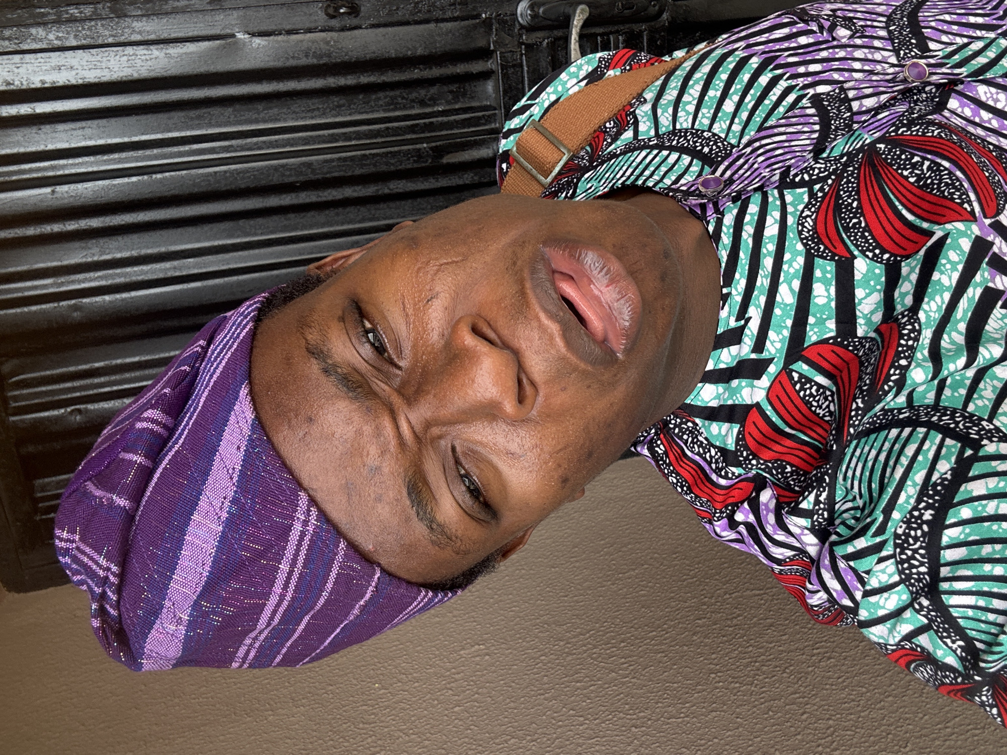 The image depicts a person wearing a purple and white striped head covering and a patterned shirt with green, red, purple, and black designs. A brown strap is visible over their shoulder. The background appears to be a dark, corrugated surface. The location is Unknown, Unknown.