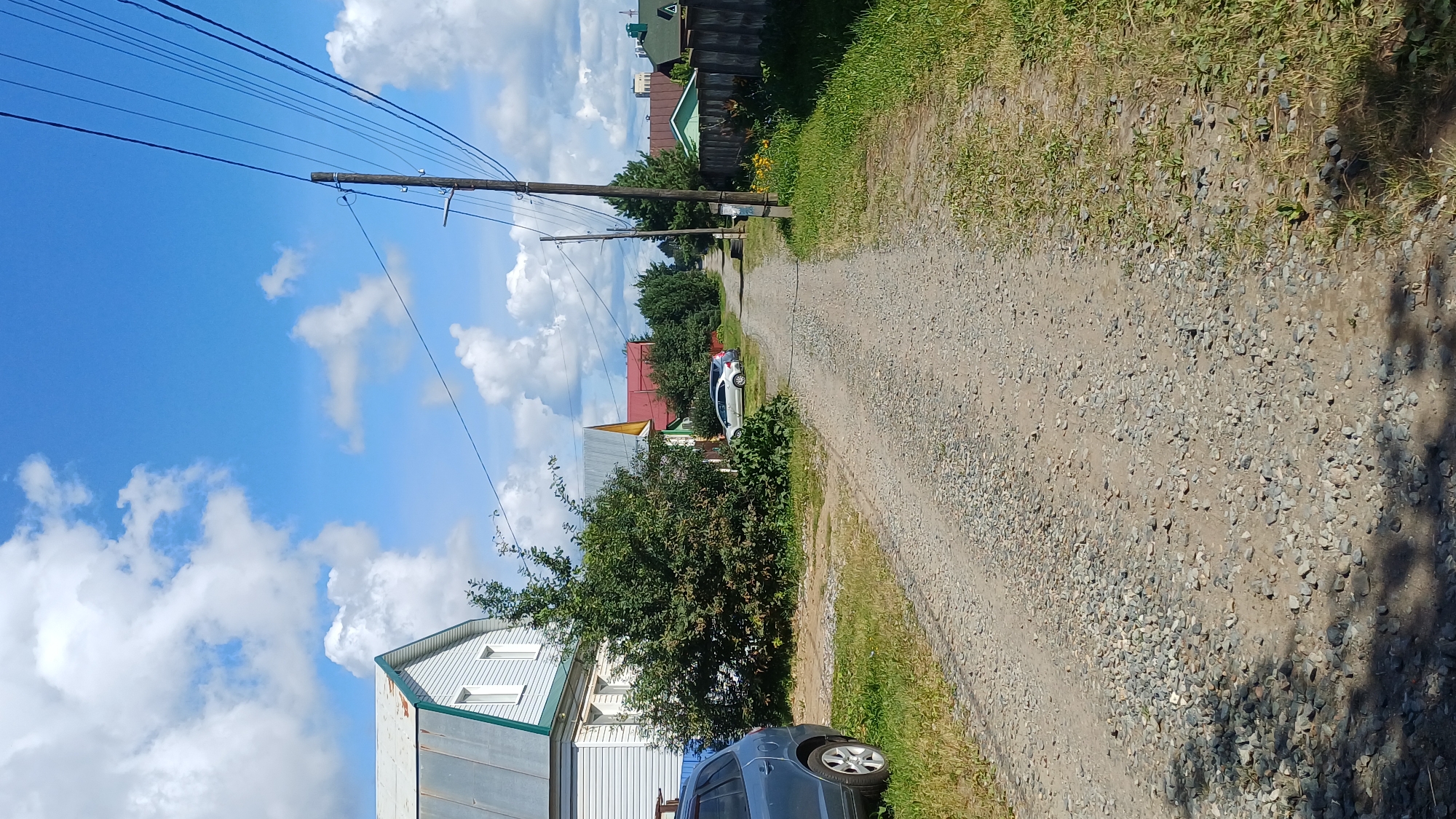 The image depicts a rural road in Izhevsk, Russia, on a sunny day with scattered clouds. The road is unpaved and covered in gravel. On the left side of the road, there is a light blue SUV parked near a light blue and white house with a metal roof. Adjacent to the house is a dense green shrub. Further down the road, on the right side, a wooden fence runs along the property line, bordering a grassy area with some yellow flowers. Several houses with colorful roofs are visible in the background, nestled among trees. Utility poles with numerous overhead wires line the right side of the road, extending into the distance. The overall scene suggests a quiet, residential area within Izhevsk.