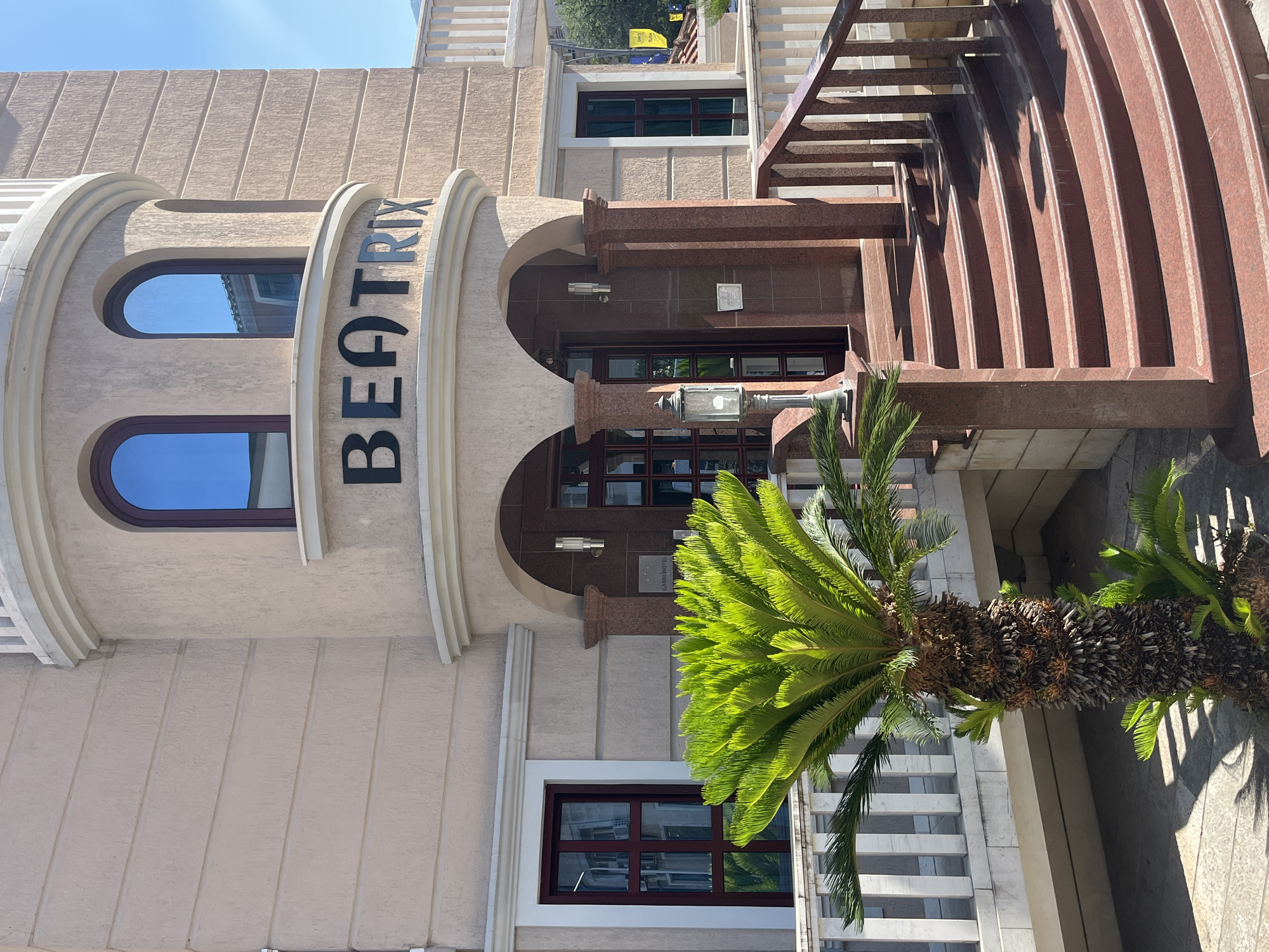 The image shows the exterior of a building named "BEATRIX" in Budva, Montenegro. The building has a beige facade with horizontal lines and a rounded corner featuring two arched windows with dark frames and reflective glass. The name "BEATRIX" is displayed in large black, sans-serif letters above the entrance. The entrance is accessed by a curved staircase with red granite steps and a matching railing. A tall palm tree with a rough trunk stands to the left of the stairs, in front of a balcony with white railings. Another smaller palm plant is visible at the base of the larger one. The surrounding environment appears sunny, with a clear blue sky visible. There are no people or animals in the frame. The overall impression is of a well-maintained building in a warm climate.