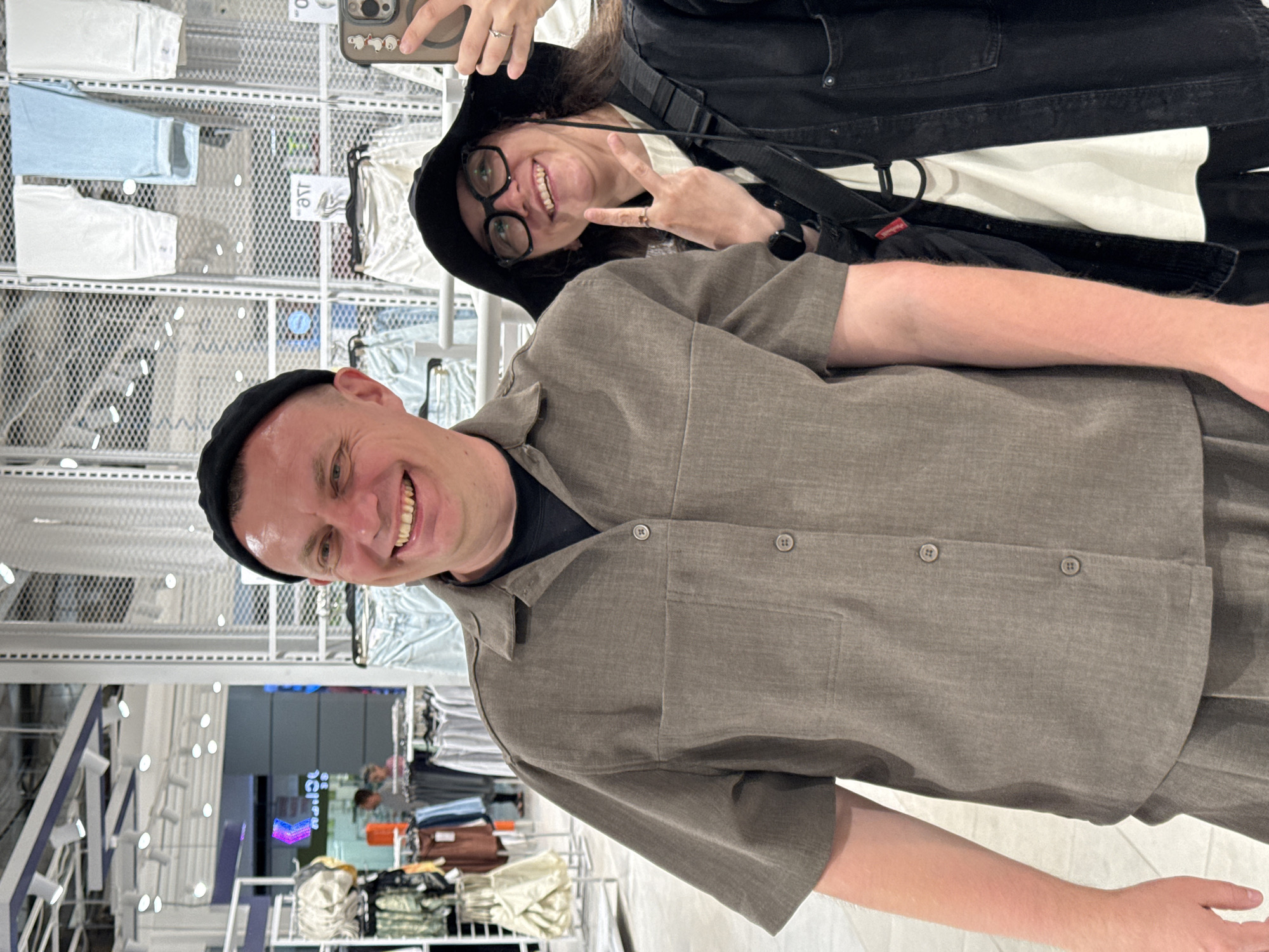 Two individuals, a man and a woman, are taking a selfie in what appears to be a retail clothing store. The man, on the left, is wearing a dark beanie and a brown short-sleeved collared shirt. The woman, on the right, is wearing a black bucket hat and glasses, and a black jacket over a white shirt. She is holding up a phone to take the picture and making a peace sign with her fingers. Behind them, clothing racks with various garments are visible, suggesting the location is a department store or fashion boutique. The background is somewhat blurred, but displays typical store lighting and shelving units. No specific geographical markers to identify Izhevsk, Russia, are present in the image.
