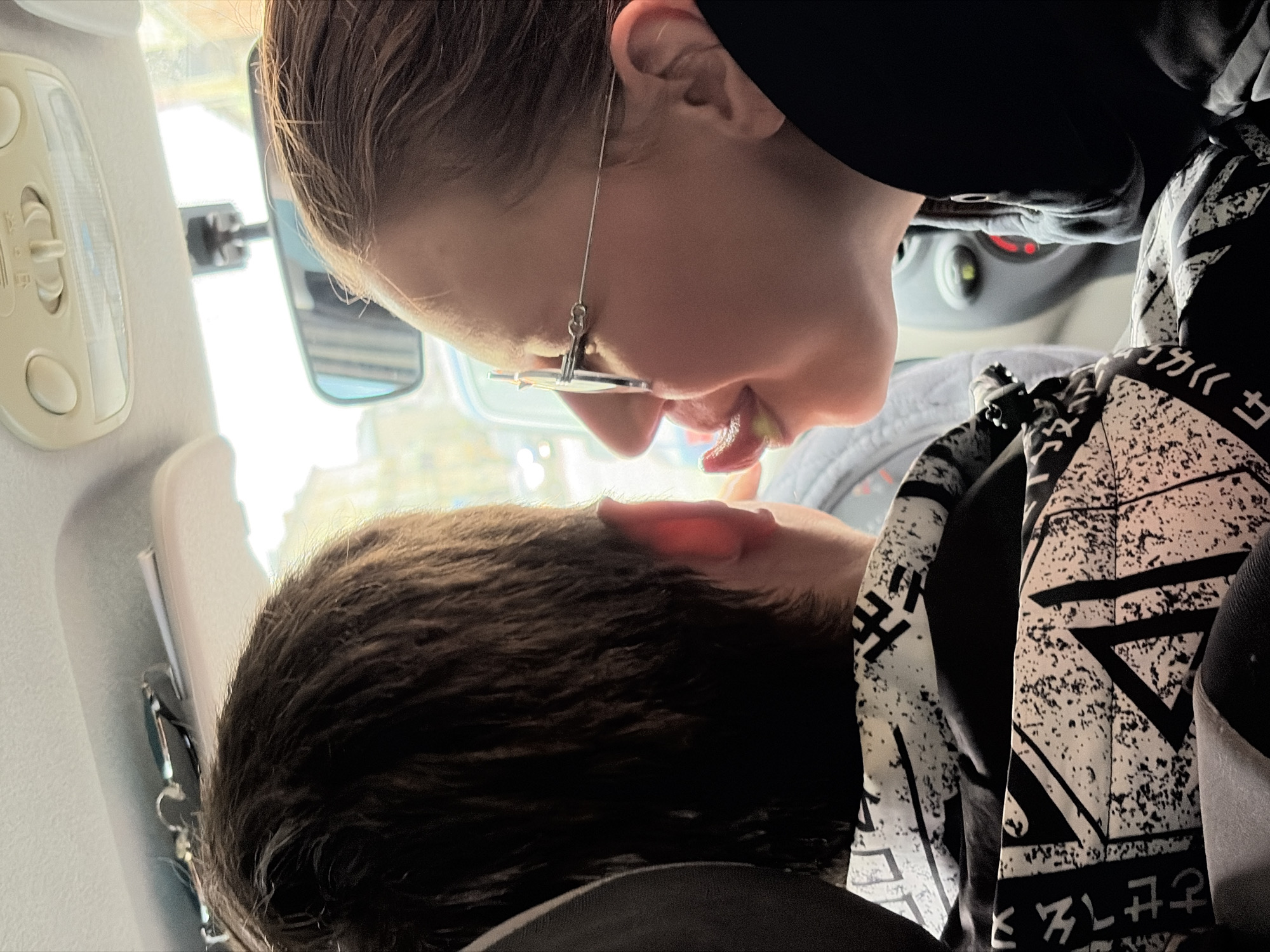This media file captures an intimate moment between two individuals inside a car cabin, viewed from an unconventional low angle looking upwards, with the image itself being rotated.
The main subject, positioned in the upper part of the frame, is a person leaning downwards. They are seen in profile from the chin to the top of the head, wearing distinctive thin, wire-frame glasses that feature a chain-like embellishment near the ear. Their mouth is slightly open, revealing part of the tongue, indicative of a kiss or a playful interaction. This individual is dressed in a dark hooded garment.
The second individual is partially visible in the lower portion of the frame, showing the back of their head with dark, wavy hair and an ear, facing upwards towards the first person. They are wearing a unique black and white patterned garment adorned with geometric shapes and characters that resemble East Asian script, specifically "ともせえそヲ" (tomoseeso wo) on the visible fabric.
The setting is clearly the interior of a car, with elements such as a rearview mirror, an overhead light console, and illuminated red indicators on the dashboard visible in the background. Bright daylight streams through the car windows, suggesting the time of day. The overall impression is one of affection and closeness. The location context for this image is Izhevsk, Russia.