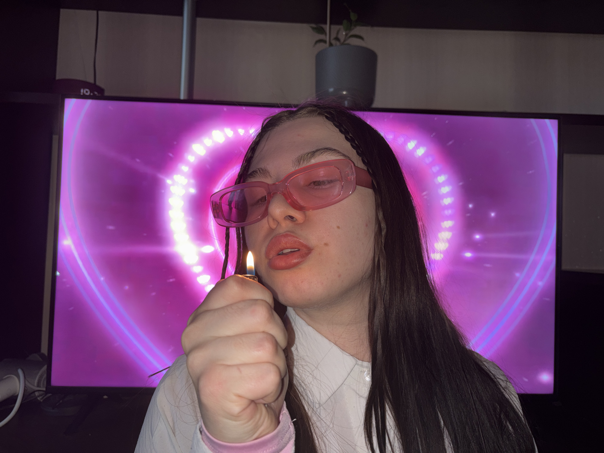 A young adult woman with long brown hair, wearing pink sunglasses and a white shirt with pink trim on the sleeves, holds a lit lighter in her right hand near her face. Her lips are pursed. Behind her is a large screen displaying a pink heart-shaped design. Above the screen are a potted plant and other objects. The location is likely indoors. There is no evidence the location is in Perm, Russia.
