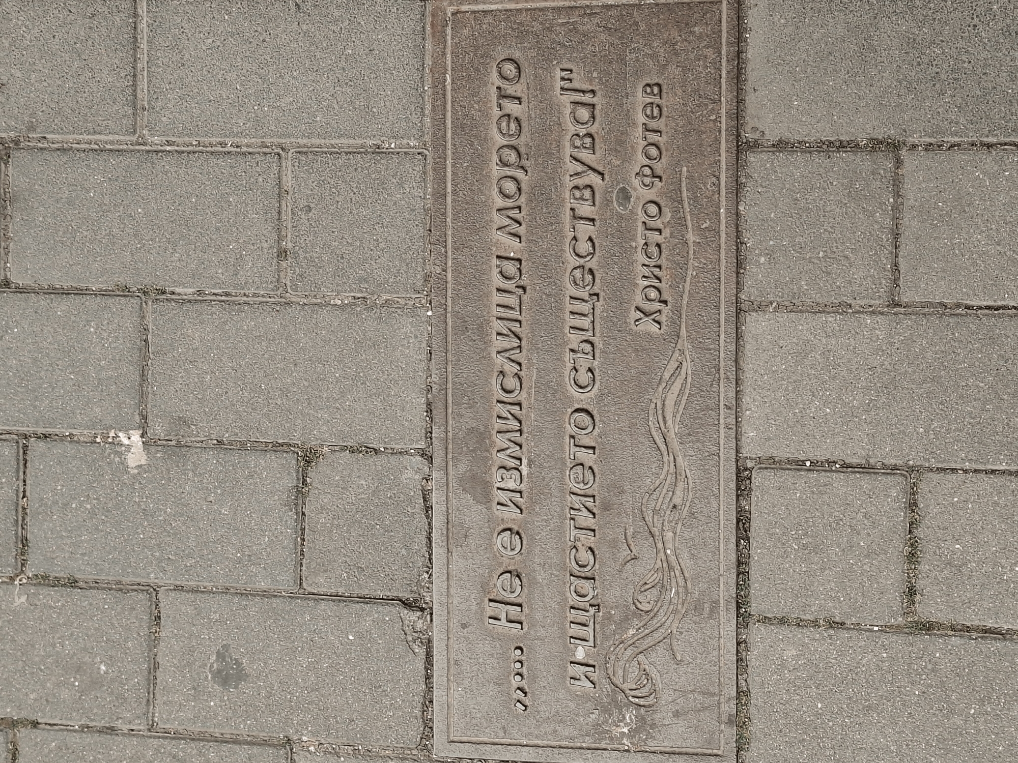 The image displays a rectangular plaque embedded in a paved outdoor surface, likely a sidewalk or pedestrian area in Burgas, Bulgaria. The paving consists of numerous light grey, speckled concrete blocks with distinct joints. The plaque itself is a darker, metallic or cast concrete slab, oriented vertically, featuring embossed text and a decorative design.
The text on the plaque is in Bulgarian and reads:
"...Не е измислица морето" (Not an invention is the sea)
"И щастието съществува!" (And happiness exists!)
Below this quote, the name "Христо Фотев" (Hristo Fotev) is embossed, identifying the poet who authored the lines.
To the right of the text, an intricate, wavy design is cast into the plaque, resembling flowing water or stylized natural elements. The plaque appears to be a public art installation or a memorial, honoring the poet and sharing his words. The scene is illuminated by ambient daylight, with no strong shadows, suggesting it could be an overcast day or midday. The surfaces show some signs of wear and dirt, indicating its integration into the urban environment.