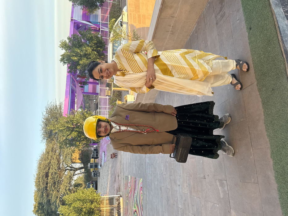 Two women stand side-by-side outdoors. The woman on top wears a yellow and white patterned outfit with a cream-colored shawl and sandals. The woman below sports a yellow helmet, a brown blazer, a long black skirt, sneakers, and holds a small, dark bag. She also wears a beaded necklace. Behind them, trees and a partially visible purple structure add to the background. The area seems to be paved and possibly near some event staging.