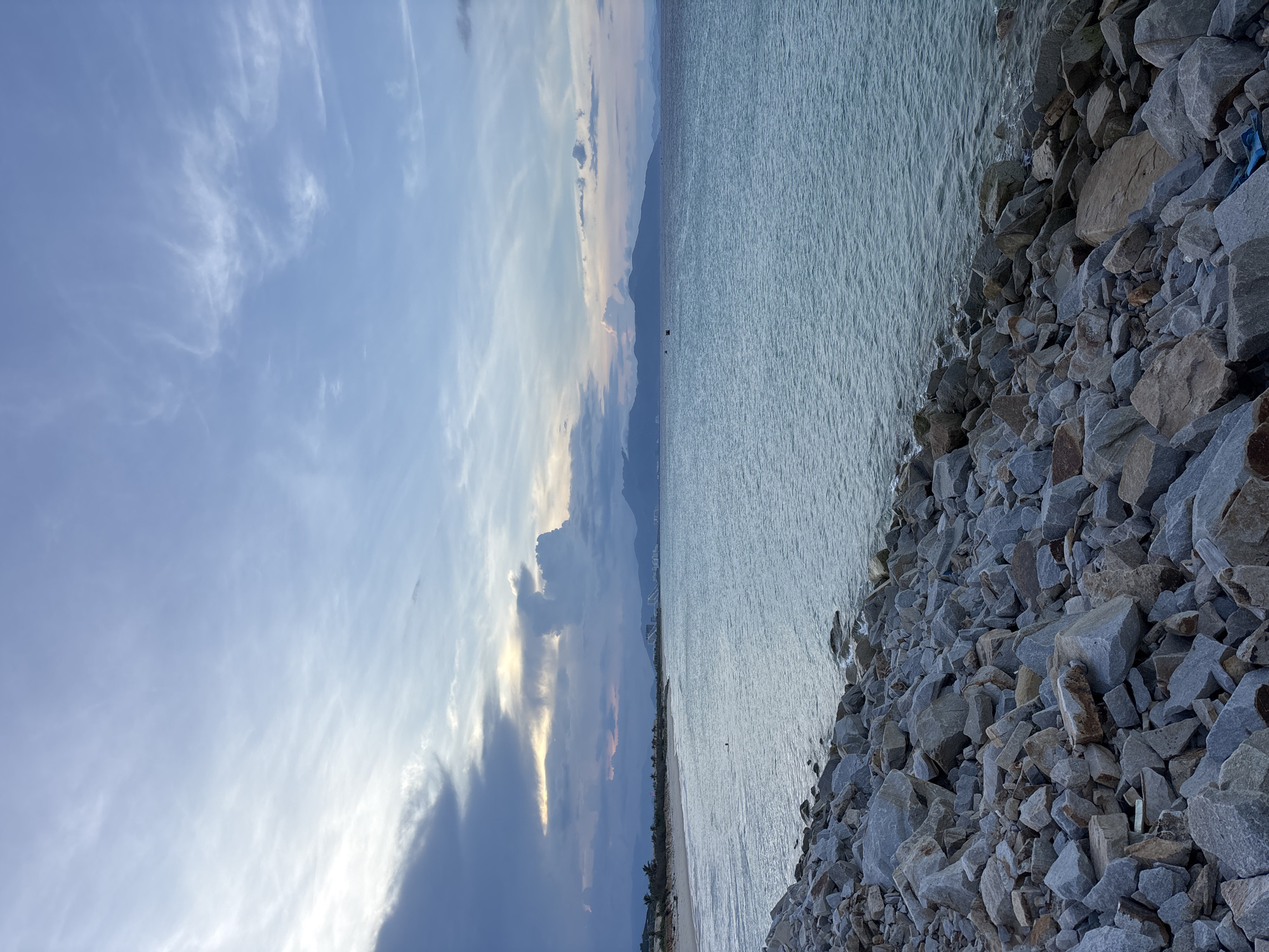 The photograph depicts a coastal scene in Bac Cam Ranh Ward, Vietnam, featuring a sky with blue hues and scattered clouds, a body of water, and a rocky shoreline. The sky transitions from light blue to darker tones with cloud formations visible. The water shows a subtle wave pattern. The shoreline is composed of medium-sized rocks. In the background, a faint outline of land and structures suggests the presence of a city or populated area.