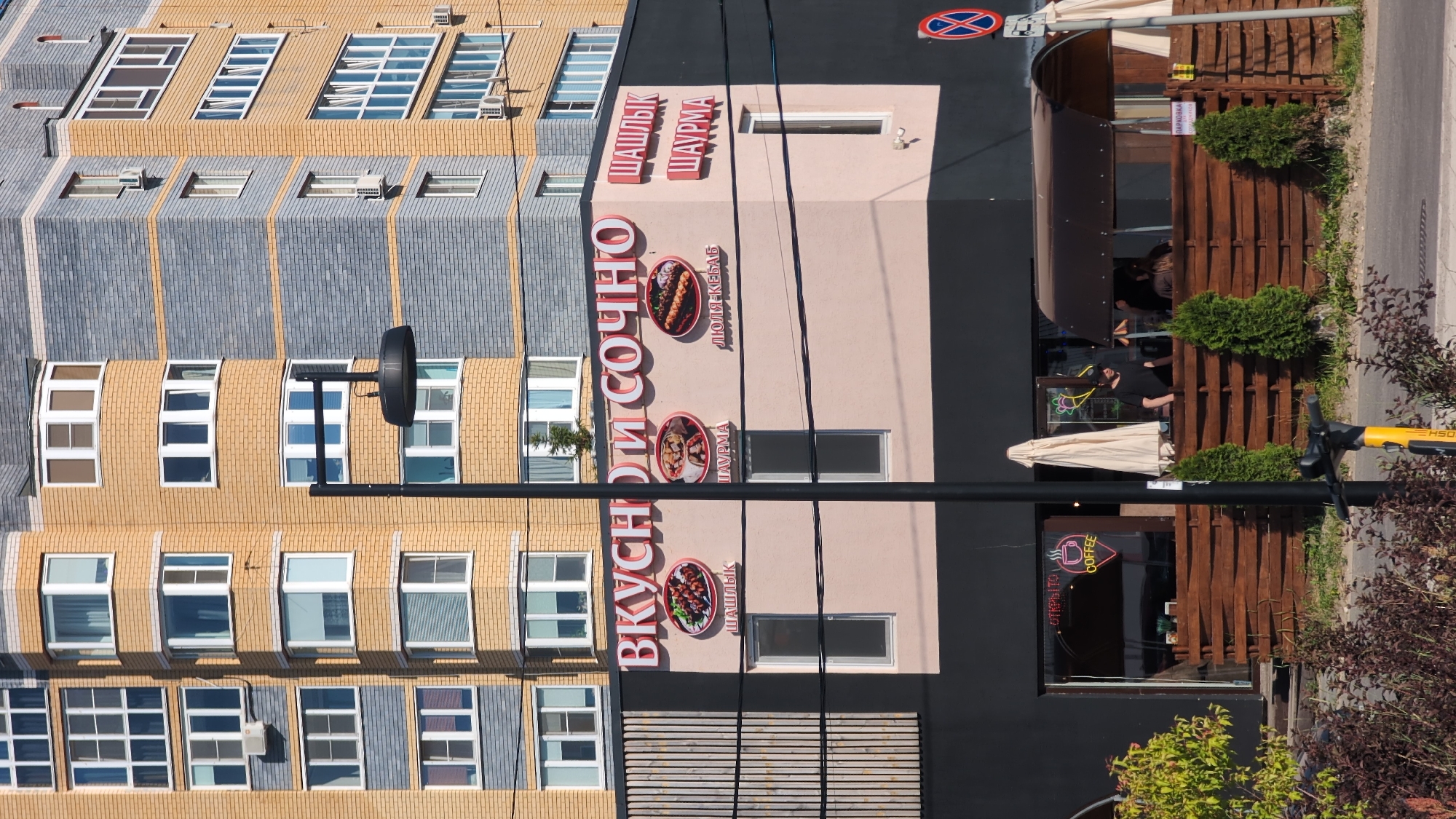 This is a medium-angle, vertical shot of a building facade in Bor, Russia. The building appears to be a commercial establishment with multiple businesses. The main focus is on the signage and facade of a restaurant that advertises "ВКУСНО И СОЧНО" (Tasty and Juicy) with images of shashlik and shawarma. Another sign reads "ШАШЛЫК" and "ШАУРМА". Below, a sign with a coffee cup logo reads "COFFEE". A wooden fence with some greenery and plants surrounds an outdoor seating area. A person in a black shirt is visible inside the establishment. A yellow electric scooter is parked in the foreground. The building has a modern design with contrasting brickwork and a dark colored upper section. The sky is not visible, but the lighting suggests it is daytime.