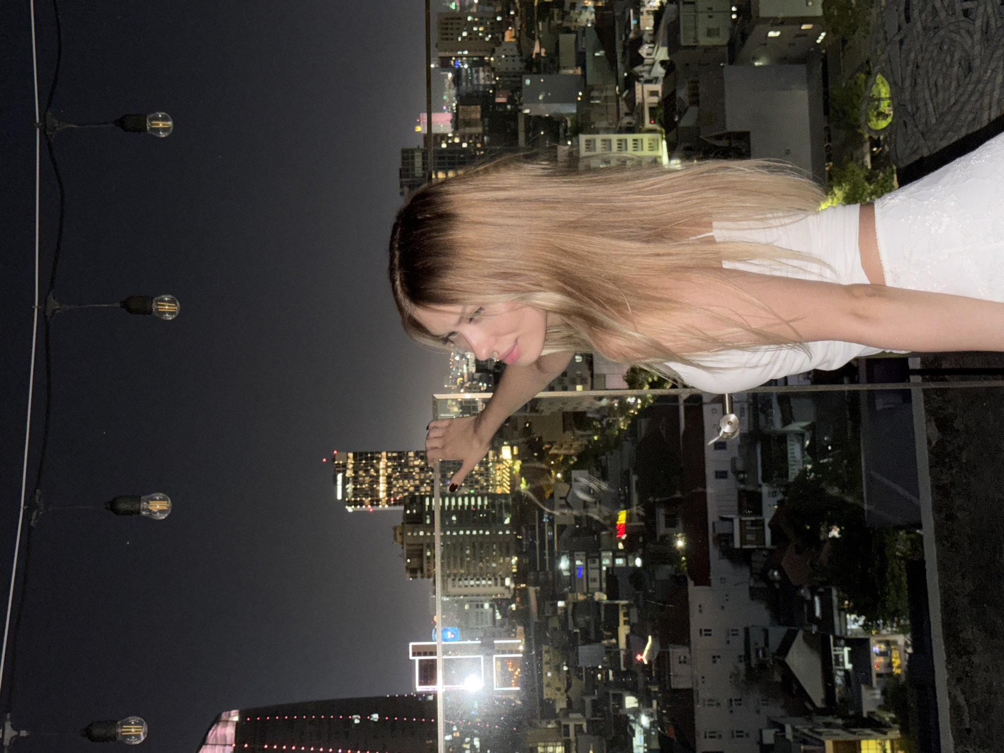 A young woman with blonde hair stands on a rooftop overlooking a city at night. She is wearing a white tank top and a white skirt. Her right arm is extended, pointing towards the city skyline. The city below is illuminated by numerous lights from buildings and streetlights. In the background, there are several skyscrapers and smaller buildings. Overhead, string lights with exposed bulbs hang across the scene. The overall impression is one of a vibrant urban environment at night. The location is the city of Đà Nẵng, Vietnam, likely from a high vantage point such as a rooftop bar or restaurant.