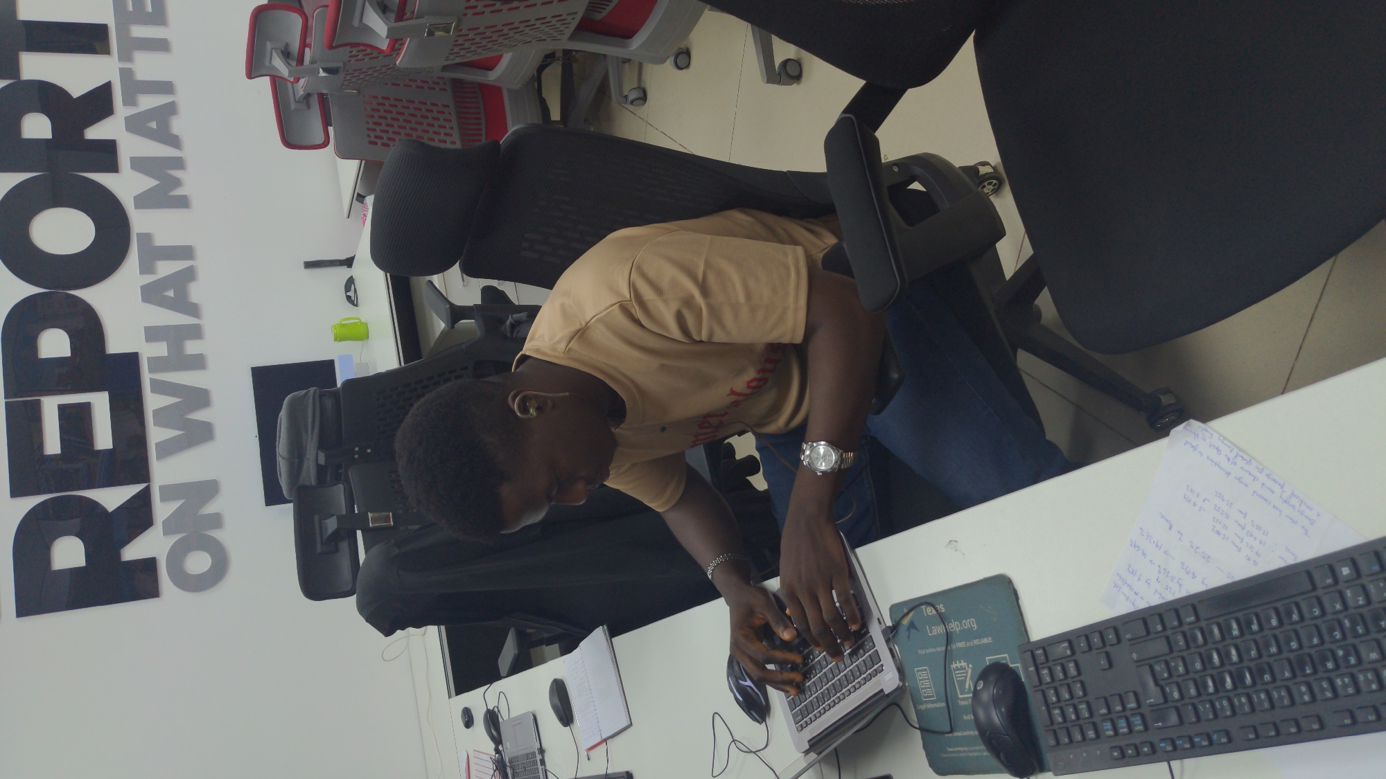 This image captures a man working on a laptop in a modern office environment in Abuja, Nigeria. The man, who has dark skin and short dark hair, is shown from a slightly elevated perspective, leaning forward over his laptop as he types. He is dressed in a light brown short-sleeved t-shirt and blue jeans, with a silver watch on his left wrist and an earbud visible in his right ear.
He is seated at a white desk in a black office chair with a mesh back. His workspace includes a silver laptop, a black wired mouse positioned on a mousepad, and an external black keyboard. The mousepad displays the text "Texas LawHelp.org" and "Your guide to FREE and RELIABLE legal information". A white sheet of paper next to the keyboard contains handwritten notes with various numbers and percentages like "17.20%", "31.31%", "4.17%", and English text that appears to be "The user has commented on your description to provide". Several notebooks, one open and one closed, are also on the desk.
In the background, a white wall features large, bold text in black and grey spelling "REPORT ON WHAT MATTERS". Other office chairs, some red and some grey, are visible, indicating a shared office space. The scene portrays an individual engaged in focused work within a well-lit, contemporary office setting.
