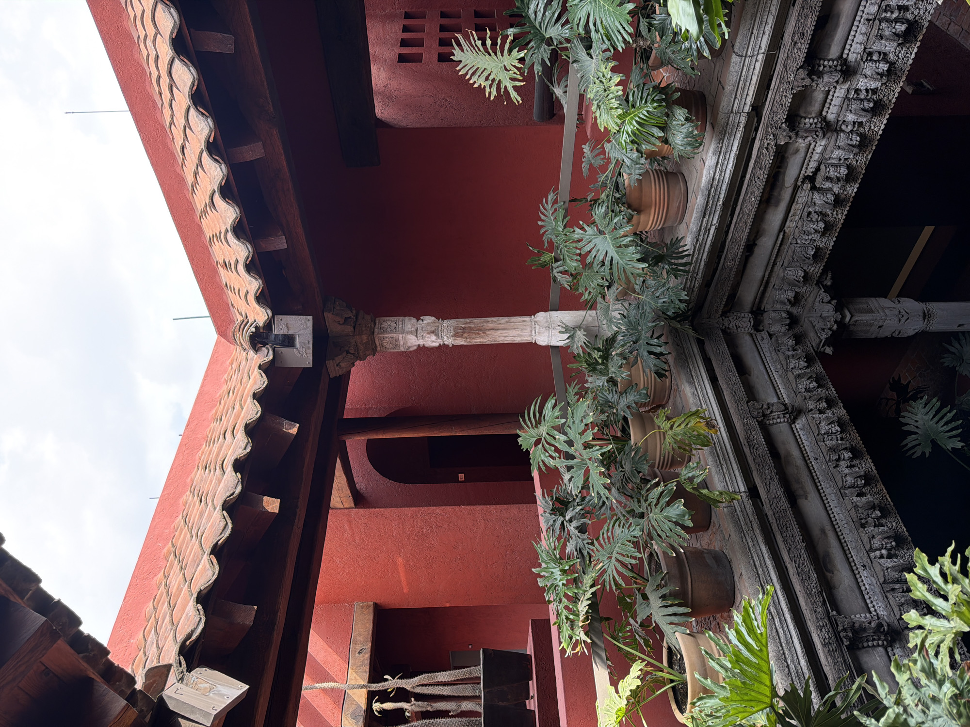The image captures an upward view into the courtyard of a building in Puebla, Mexico. The architecture is characterized by red-painted walls and intricate woodwork on the building's trim and support structures. Two main architectural elements are visible on either side of the frame: one with a clay-tiled roof edge and the other with ornately carved dark wooden trim. A decorative, light-colored pillar supports the structure. Potted plants with large green leaves are suspended along the walls. The sky is visible through the open top of the courtyard.
