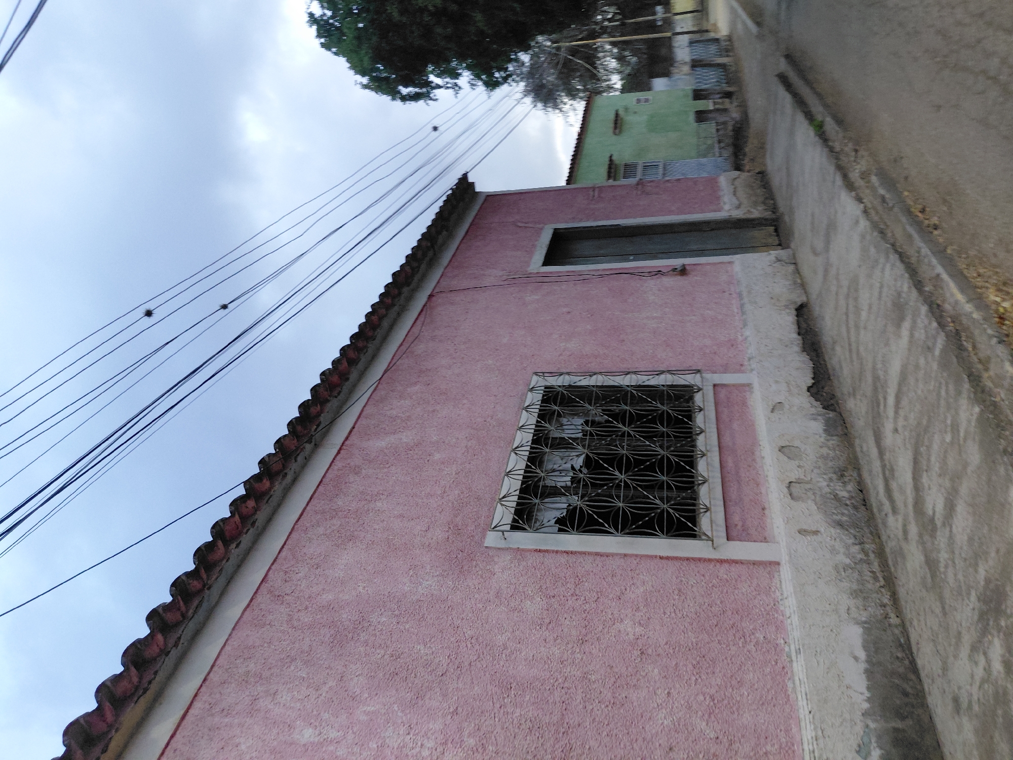 The image presents a residential street view in Santa Ana, Venezuela, under an overcast sky. A single-story structure with a textured, faded pink stucco facade dominates the left and central foreground. Its roof is constructed from terracotta-colored corrugated tiles, exhibiting wear and an overhang. A rectangular window, framed in white, is set into the pink wall and protected by a dark metal security grate featuring interwoven circular patterns. To the right of the window, a recessed dark wooden door, also with a white frame, provides access. The lower portion of the building displays a white-painted base that is significantly chipped and degraded, exposing a darker grey substrate. A grey concrete sidewalk, showing signs of wear and minor cracking, runs along the base of the pink building.
Multiple black utility wires extend diagonally across the upper section of the frame, originating from the left and ascending towards the upper right. Two small, dark, irregular, globular objects, consistent with bird nests or entangled debris, are suspended from distinct wires in the upper left quadrant.
In the mid-ground, a second single-story building with a light green facade is partially visible to the right of the pink structure. This building also features a tiled roof and a barred window or door. A low, light-colored fence with vertical bars is situated in front of the green house. A large tree with dense, dark green foliage is present on the far right, partially obscured by the utility lines. The street appears unpaved or composed of dirt, with a narrow concrete or paved strip running adjacent to the sidewalk of the pink building, possibly functioning as a gutter. The sky is uniformly cloudy, characterized by various shades of grey and white, indicating diffused light. No people are discernible within the scene.