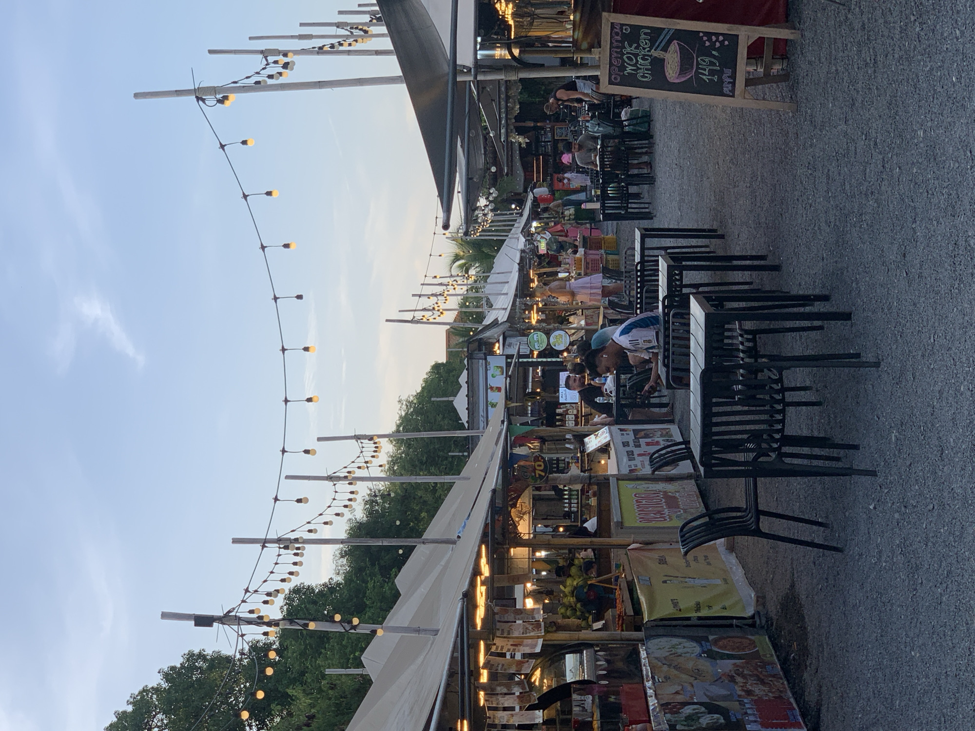 This is a photograph taken in the late afternoon or early evening at a vibrant outdoor market in Ko Samui District, Thailand. The market is characterized by rows of stalls with covered awnings, illuminated by strings of decorative lights. Several food stalls are visible, displaying menus and food items. People are seen browsing, eating at tables, and working at the stalls. The ground is covered in gravel. In the foreground, there are several empty tables and chairs set up for diners. In the background, more stalls and people can be observed, along with lush green trees and a light blue sky. A chalkboard sign in the right side of the image advertises "open now wok chicken" with a price of 149. The overall atmosphere suggests a lively and bustling marketplace.