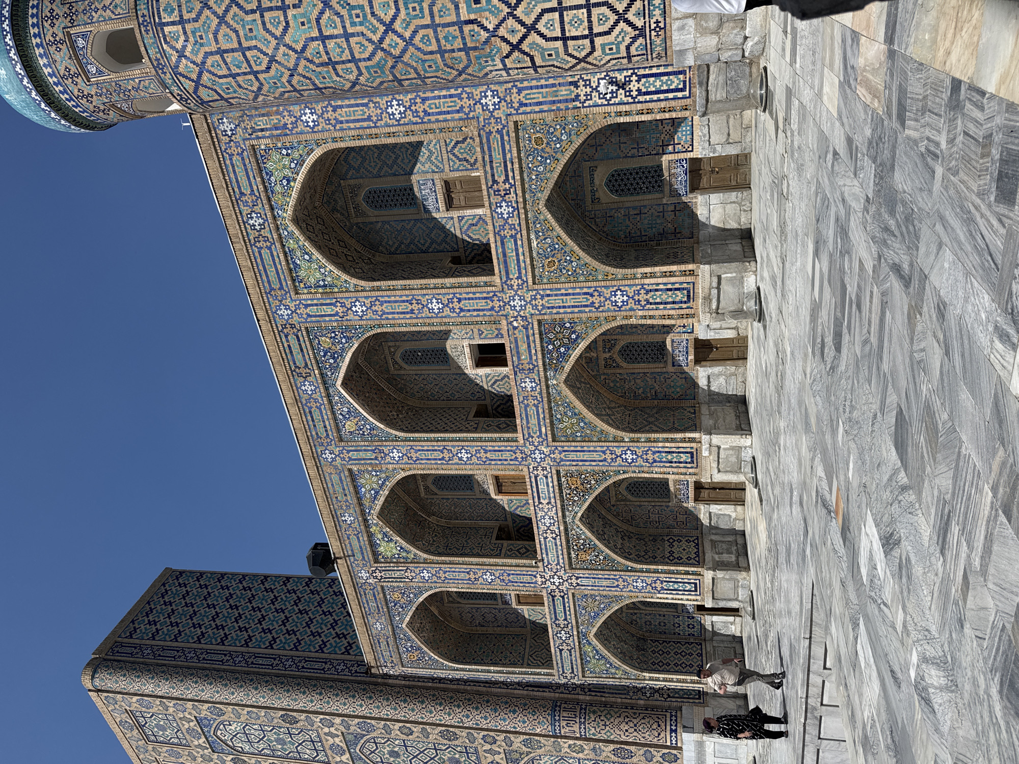 The image shows a building in Samarkand City, Uzbekistan, likely part of a historical complex. The building features intricate tile work in blue, turquoise, and beige hues. The architecture includes a series of arched openings or niches on the upper levels, with smaller windows visible within some of the niches. The ground level is paved with large, gray stone slabs. Two people are standing on the pavement. The sky is clear and blue. The overall impression is of a well-preserved historical site with detailed ornamentation.
