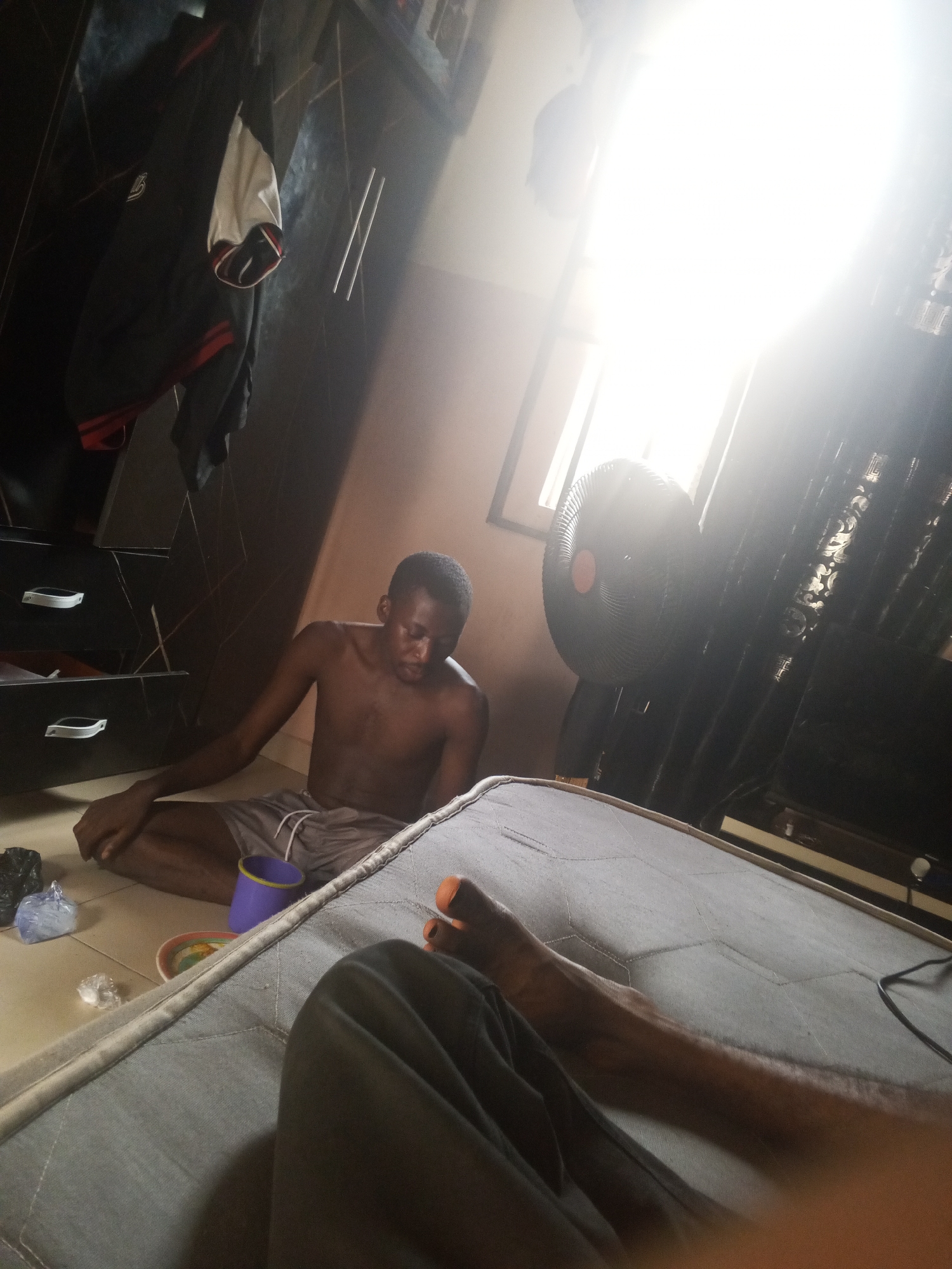 In an indoor setting, a young Black male, shirtless and wearing gray shorts, is seated on the floor. He appears to be looking down and slightly to his right. To his right, a black oscillating fan is positioned near a window with bright light streaming through. Behind him, a dark dresser with multiple drawers is visible, and a jacket hangs on the left side. In the foreground, a mattress is partially visible, with another person's legs and feet, wearing orange-painted toenails, positioned on it. Various objects, including plastic bags, a cup, and a bowl, are scattered on the floor near the male. The room's walls are a light pinkish-brown color.