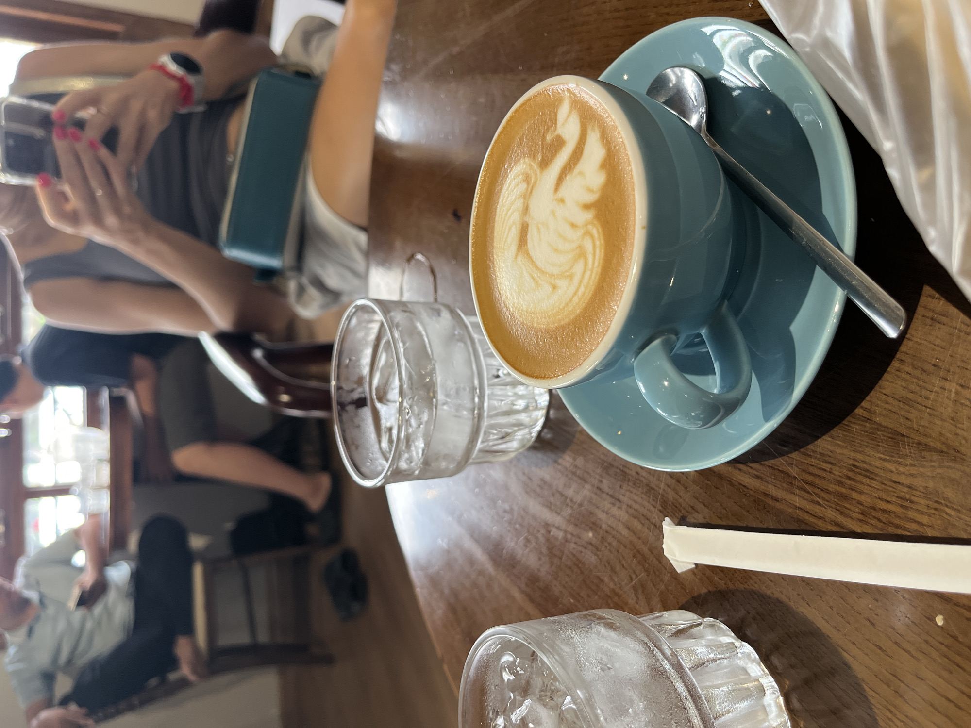 The image shows a table in what appears to be a cafe, potentially in Nha Trang Ward, Vietnam. A blue cup of latte with latte art sits on a matching saucer with a spoon. Two glasses, one apparently containing water, and a paper-wrapped straw are nearby. A person in the background is holding a phone, possibly taking a picture. Other blurred figures are also visible in the background. The table is wooden and shows signs of wear.
