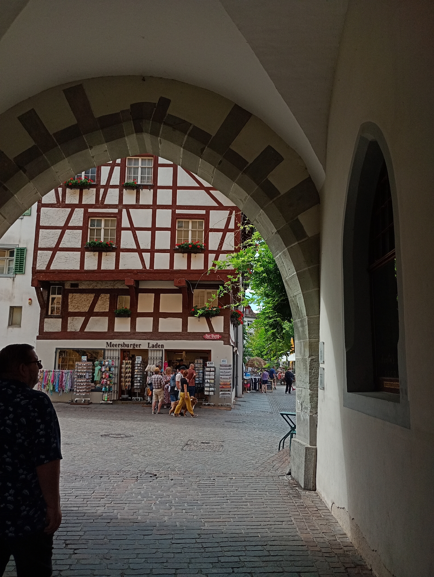 The image shows an archway in the city of Meersburg, Germany, framing a view of a cobblestone street lined with historic buildings. In the foreground, a man wearing a dark blue patterned shirt stands with his back to the camera. Through the arch, a half-timbered building with red trim and white infill is visible. Below the building, "Meersburger Laden" is written above a shop entrance. Several people are walking and standing on the street, some looking at merchandise displayed outside the shop. A green metal chair is visible to the right of the street. Lush green trees overhang the street in the background. The cobblestone pavement is worn and uneven.