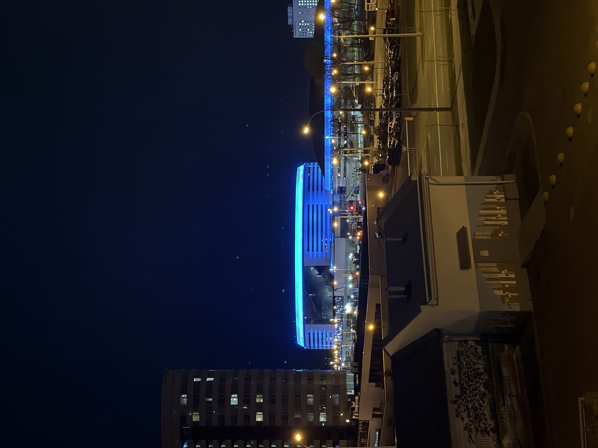 The image captures a vibrant urban night scene, presented with a 90-degree counter-clockwise rotation from its natural landscape orientation. When viewed correctly, a dark, clear night sky fills the upper portion, devoid of clouds or visible stars, indicating a clear night.
The central focus is a large, modern building distinguished by striking, vibrant blue illumination along its upper structures and facade, giving it a futuristic aesthetic. This edifice, possibly a stadium or a major event complex, dominates the mid-ground. To its left, a tall residential or office building is visible, with numerous illuminated windows creating a grid pattern of warm light against its dark facade.
In the foreground and extending to the right, a street or road is visible, bordered by a row of warm yellow-orange streetlights that cast their glow on the pavement and a line of parked cars. A traffic light showing a red signal can be observed near the prominent blue building. Further to the right, other smaller buildings and structures are discernible, including one with a light-colored facade featuring intricate, abstract patterns or a mural.
The scene is characterized by the stark contrast between the deep indigo of the night sky and the various sources of artificial light. The blue illumination of the central building provides a cool, distinctive hue, while the streetlights offer a warm glow. No people or explicit activities are visible, but the presence of vehicles and illuminated buildings signifies an active urban environment. No legible text is discernible within the image.