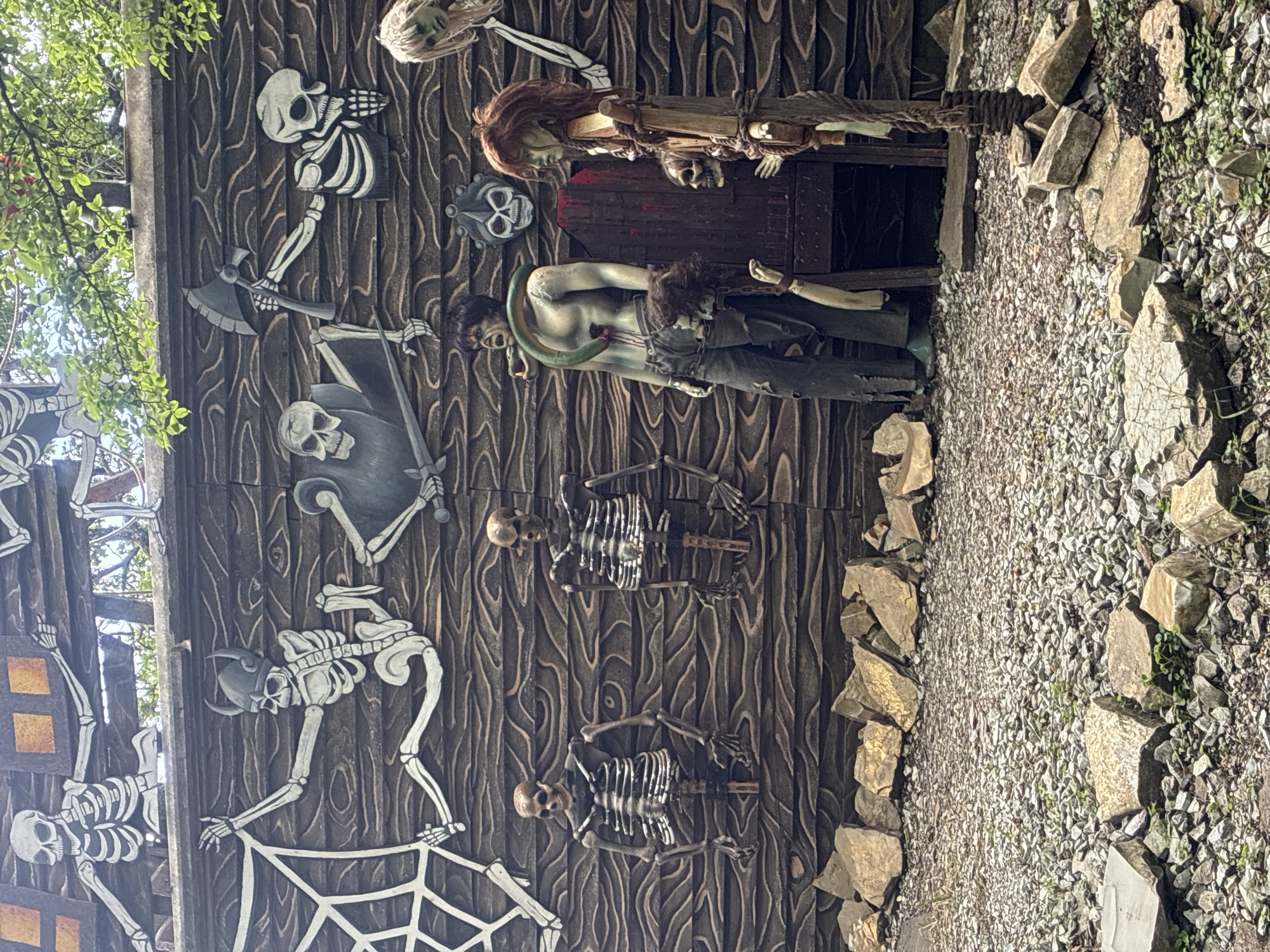 The image depicts an outdoor scene with a wooden wall decorated with various macabre figures and a rocky ground. The wall features several skeletal figures, some appearing to be climbing or posed in dynamic positions. One skeleton is depicted with a horned helmet, another wields an axe, and others are engaged in or near spiderwebs. In front of the wall, there are several mannequins arranged to create a disturbing tableau. A male mannequin is positioned with a green snake wrapped around its neck and torso, and what appears to be a severed head hanging from its belt. To its right, another mannequin, seemingly female with long, disheveled hair, stands beside a wooden structure resembling a torture device or chair, adorned with a skull and rope. The ground is covered in gravel and scattered rocks of various sizes. The overall atmosphere is theatrical and designed to evoke a sense of horror or a haunted attraction. No specific location in Gelendzhik, Russia, can be identified from the visual information provided.