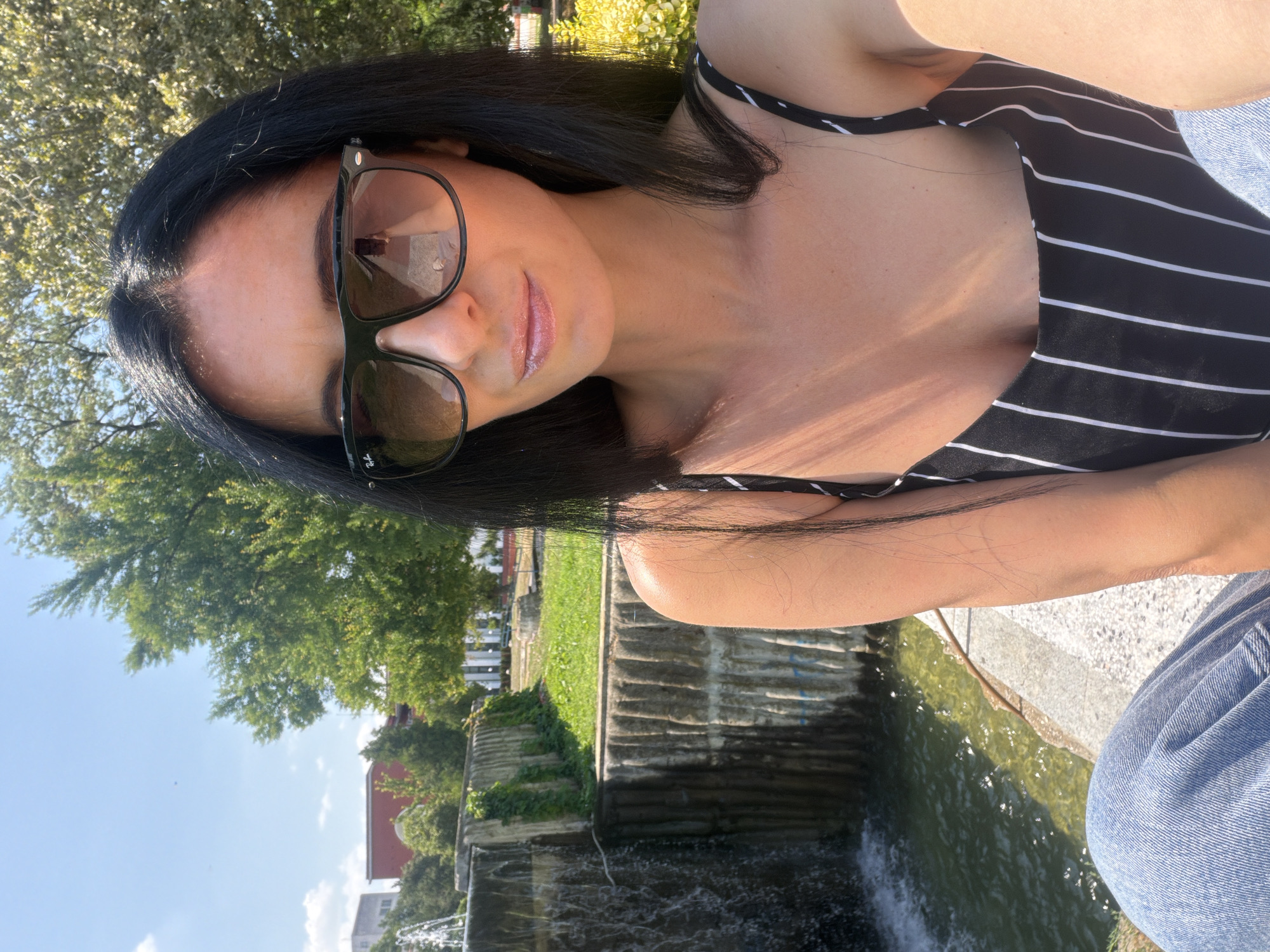 A woman with long dark hair and sunglasses is taking a selfie in front of a cascading water feature and a park area. She is wearing a black and white striped top and blue jeans. The environment appears to be a public park with trees and buildings in the background, suggesting an urban setting. The woman is seated by the edge of the water feature, which has concrete walls and some greenery. The sky is clear blue with a few scattered clouds. The overall impression is a sunny day in a recreational area. The location is identified as Pleven, Bulgaria.