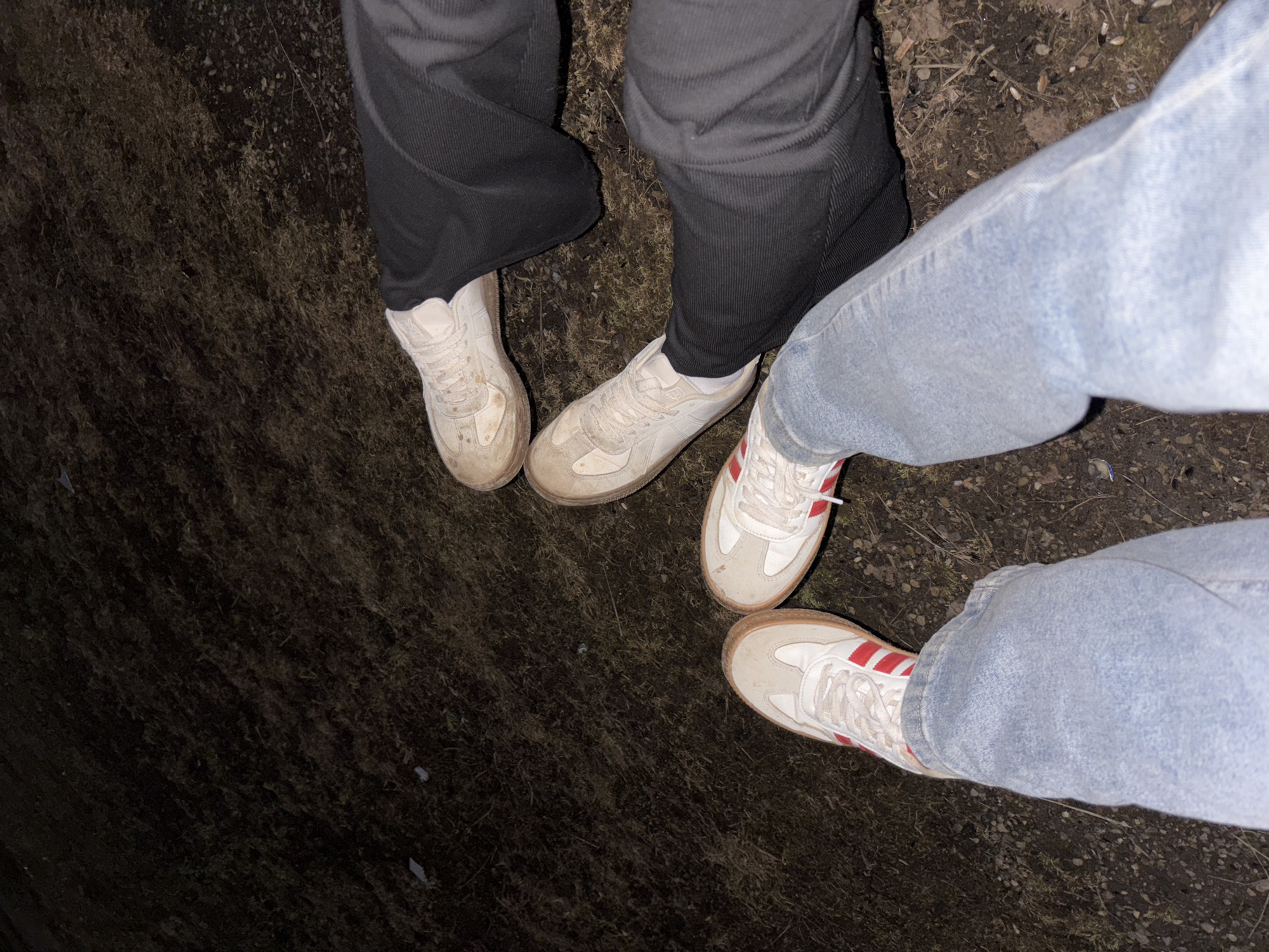 From an elevated perspective, the image captures two pairs of legs and feet standing on a dark, earthy surface, possibly soil or dirt. The legs on the left are clad in dark gray corduroy pants and off-white sneakers. The legs on the right wear light blue jeans with white sneakers that have red stripes. The surrounding environment appears dimly lit. The exact location is not discernible from the image.