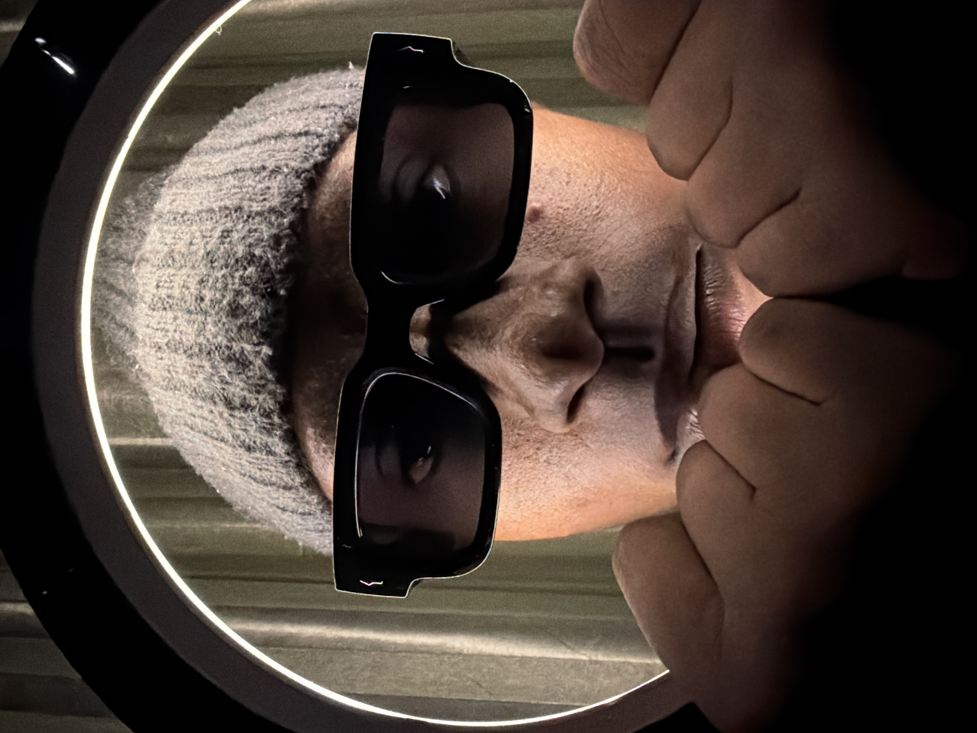 A close-up, low-angle, studio portrait of a person wearing a grey beanie and black sunglasses, with their hands cupped near their chin. The person is illuminated by a ring light, creating a bright halo effect behind them. The background consists of vertical, textured curtains in a muted green color. The image does not contain any elements that indicate a specific location in Jiangjin, China.