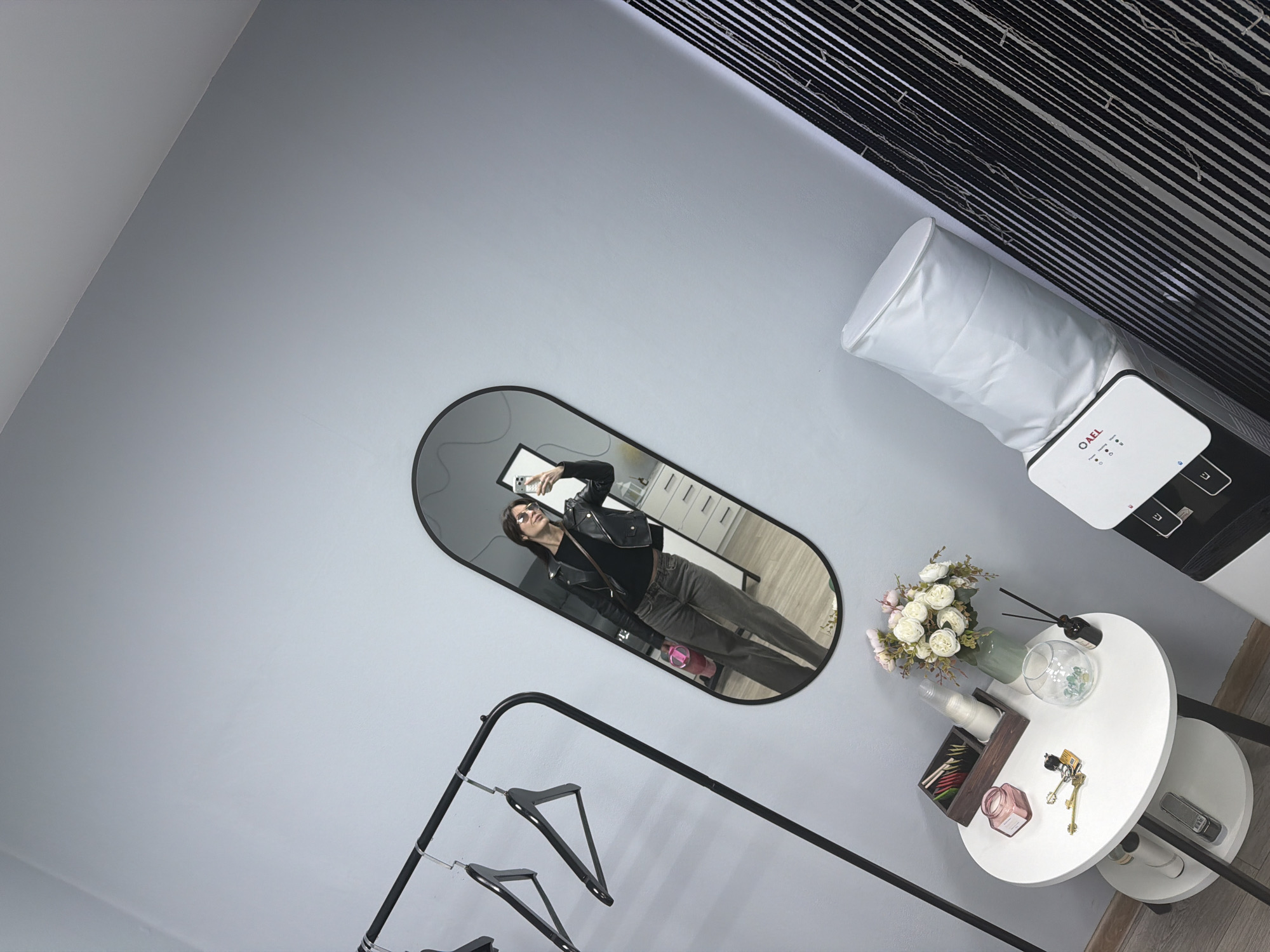 This appears to be a staged interior scene, likely in a shop or showroom, potentially in Perm, Russia. A woman takes a selfie in an oval mirror. She is wearing a black leather jacket, a dark top, and grey trousers. The mirror reflects a room with white drawers. To the right of the mirror is a white cylindrical object connected to a machine, alongside a round white table with flowers, perfume bottles, keys, and other small decorative items. To the left of the mirror is a black clothing rack with hangers. The walls are painted in a neutral grey, and the ceiling in the top right has black and white stripes.