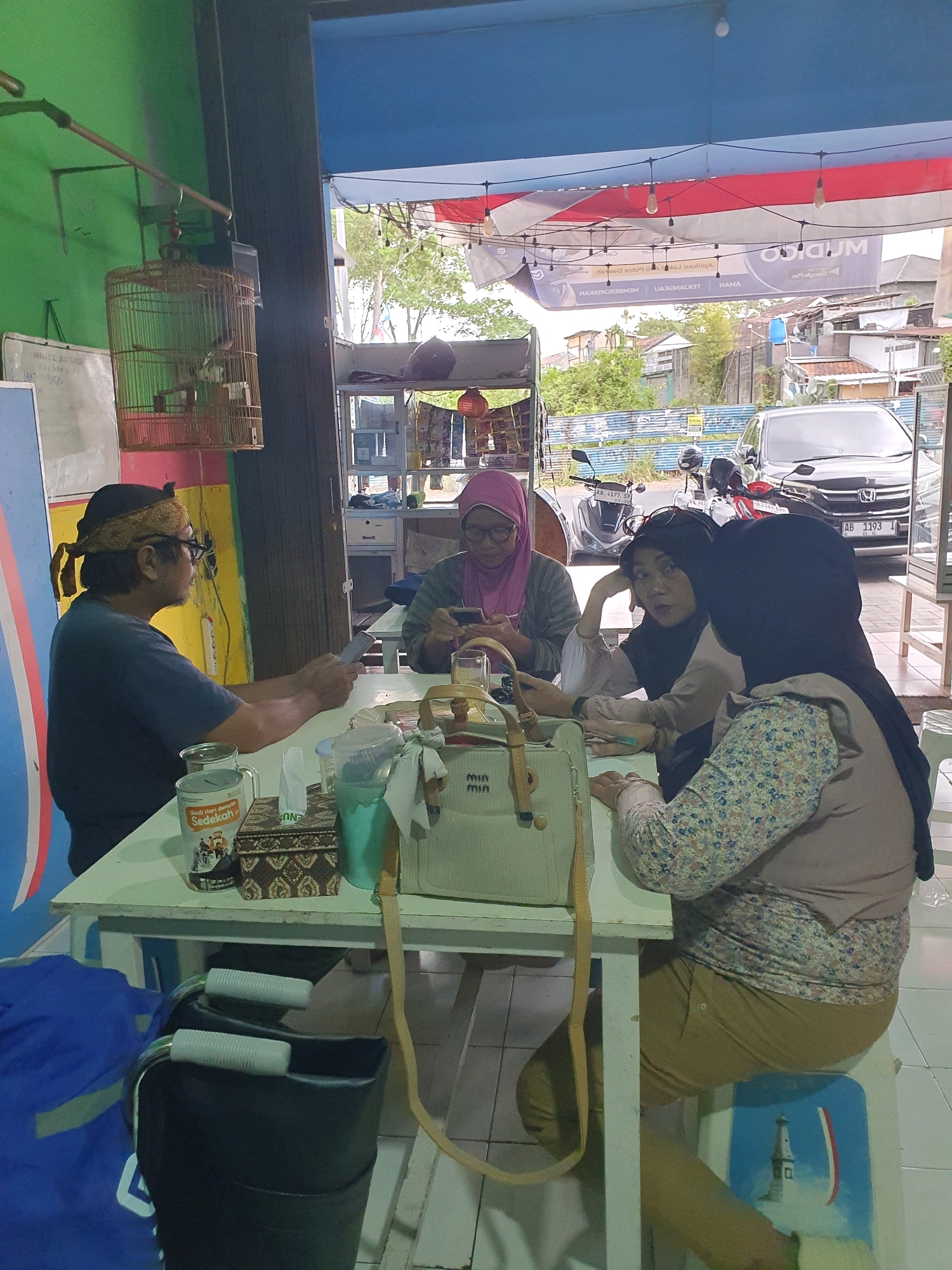In Ngaglik, Indonesia, four individuals are seated around a white table. A man on the left, wearing a dark blue shirt and a traditional headband, holds a mobile phone. To his right, a woman in a pink headscarf is also engaged with a phone. Two other women are seated opposite them; one in a light pink hijab leans on her hand, looking forward, while the other, wearing a floral patterned top and a beige hijab, looks down at the table. The table is set with drinks, a tissue box, a patterned container, and a light-colored handbag. In the background, a birdcage hangs from a metal pole. The scene appears to be an open-air eatery or shop. Outside the establishment, vehicles including a car and motorcycles are visible.