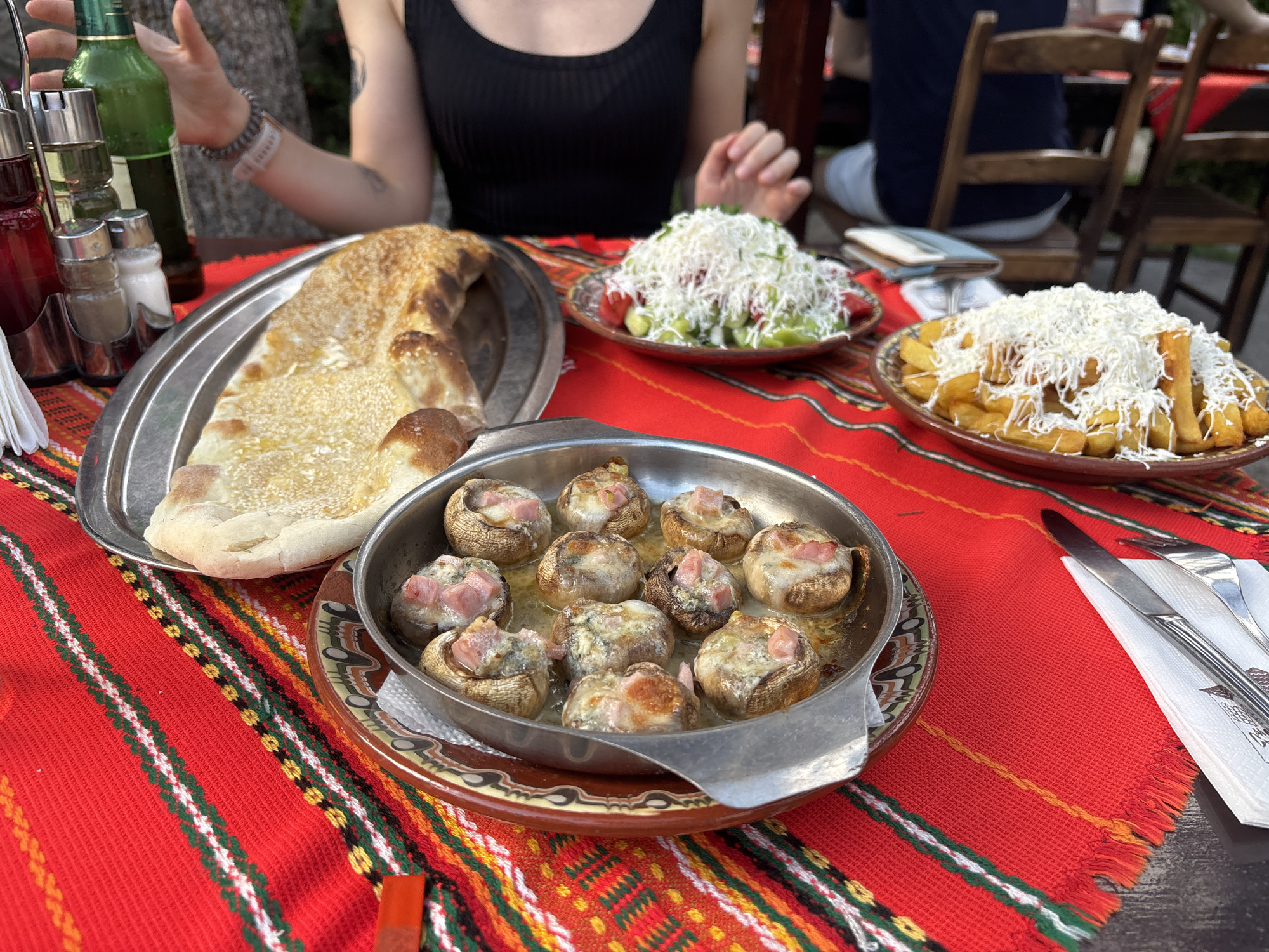 The scene captures a dining table laden with food at an outdoor establishment in Samokov, Bulgaria. In the foreground, a vibrant red tablecloth with traditional Bulgarian patterns, featuring horizontal stripes in green, white, yellow, and black, covers the table.
On the table, a diverse meal is presented. In the center foreground, a shallow, round metal pan contains approximately ten baked mushrooms, each stuffed and topped with melted cheese and small pieces of ham or bacon, resting in a clear liquid. To the left, a large, elongated flatbread with a golden, crisp crust is displayed on a silver metal tray. Further back on the left, a ceramic bowl holds a salad composed of sliced green cucumbers and red tomatoes, generously topped with grated white cheese. Mirroring this on the right, a ceramic plate piled with golden-brown French fries is also heavily garnished with grated white cheese.
A metal caddy on the far left holds several condiments: two bottles, one with dark red liquid and another with clear liquid, and a green glass bottle (likely beer). Two shakers, one with white powder (salt) and one with dark powder (pepper), are also in the caddy, alongside a stack of white paper napkins. On the far right, a knife and fork rest on a folded white paper napkin.
Two individuals are partially visible at the table. In the immediate foreground, an adult person wearing a black sleeveless top is seated, with their hands resting on the table; one hand is near the green bottle. In the background, a second person is seated with their back to the camera, wearing a dark blue top and light-colored lower garments, with wooden chairs visible around them. Green foliage is discernible in the distant background, suggesting an outdoor setting. The lighting indicates daylight.