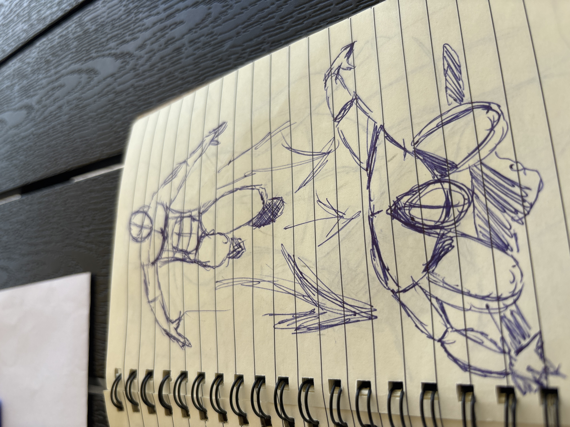The image shows a close-up of a spiral-bound notebook with a rough sketch in progress. The sketch, drawn in blue ink, depicts two figures in what appears to be a dynamic action scene, possibly a fight or a dance.
The figure on the left is more fully rendered, showing a torso with abdominal muscles indicated by cross-hatching and an arm extended forward. The head is represented by a circle with intersecting lines. The body is oriented horizontally.
The figure on the right is less defined, with more abstract lines and shapes. It appears to be larger and possibly in motion, with parts of its limbs and torso suggesting an active pose. There are several oval shapes and angular lines that could represent limbs or other elements of the figure. Some areas are shaded with scribbled lines.
The notebook paper is lined, and the sketch spans across several pages. The background of the image shows a dark, textured surface, possibly a wooden table, with the notebook resting on it. There is also a white piece of paper visible on the left edge of the frame, out of focus.
The lighting suggests it could be daytime, possibly outdoors or near a window, given the natural light illuminating the sketch and the dark background. There are no explicit details about the weather or specific emotions conveyed by the figures, as the sketch is in an early stage. The location context of Karviná, Czechia, is provided but not visually evident in the image itself. No text is visible in the image.
