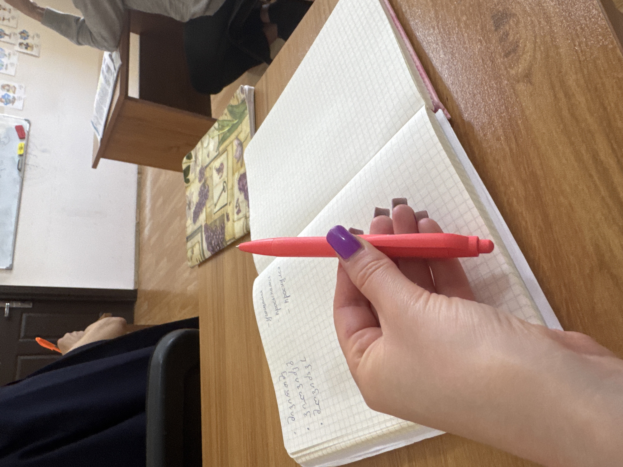 A person is writing in a notebook with a red pen. The person's hand is visible, and they have purple nail polish. The notebook has a graph paper pattern. The writing in the notebook is in both a Cyrillic and a Georgian script. A textbook with a yellow and purple pattern is visible to the left of the notebook. The setting appears to be a classroom, with a desk and a student in the background. The city and specific location are unknown.