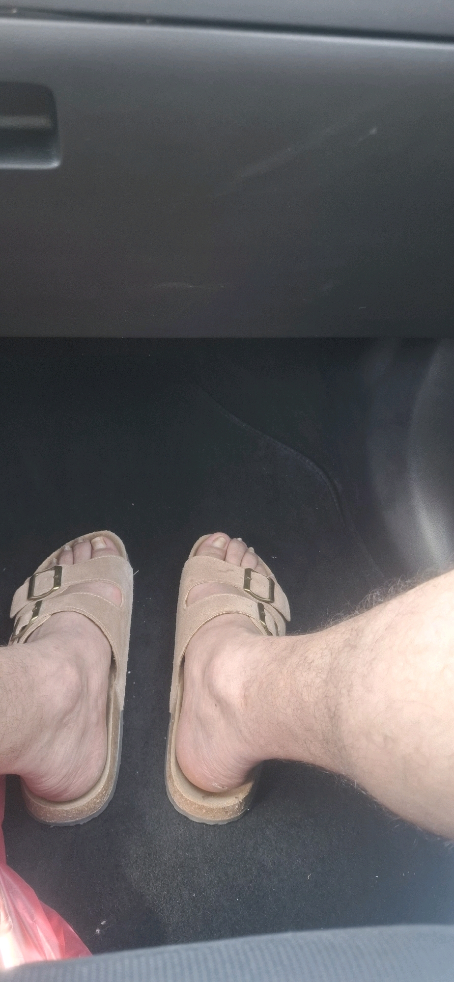 The image shows a person's feet wearing tan Birkenstock-style sandals, resting on a dark carpeted surface. The feet are bare, and one foot is slightly more visible than the other. The background appears to be the interior of a vehicle, with a dark grey dashboard above the feet. A corner of a pink plastic bag is visible in the lower left of the frame. The scene is likely indoors or inside a vehicle, and there is no information to suggest it is located in Dimona, Israel.
