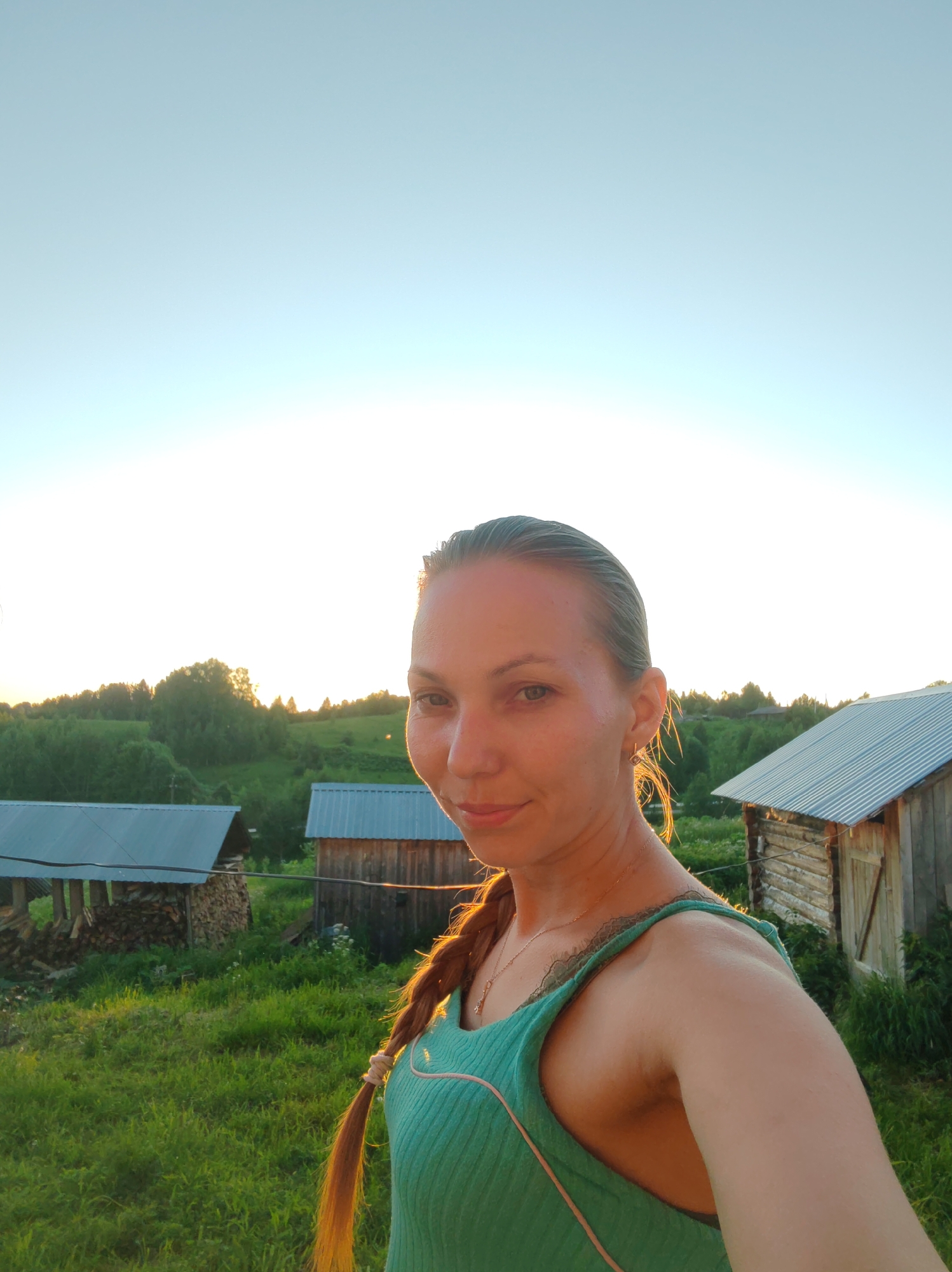 A woman with blonde hair braided and pulled to her right side, wearing a teal tank top with lace trim, poses for a selfie outdoors. She is looking at the camera with a slight smile. The background shows a green, grassy landscape with several wooden buildings with corrugated metal roofs and piles of firewood. In the distance, there are trees and a hill. The sky is a clear, pale blue, suggesting it is either early morning or late evening. The light indicates a sunset or sunrise. The image appears to be taken in the rural area of Chuchlam, Russia.