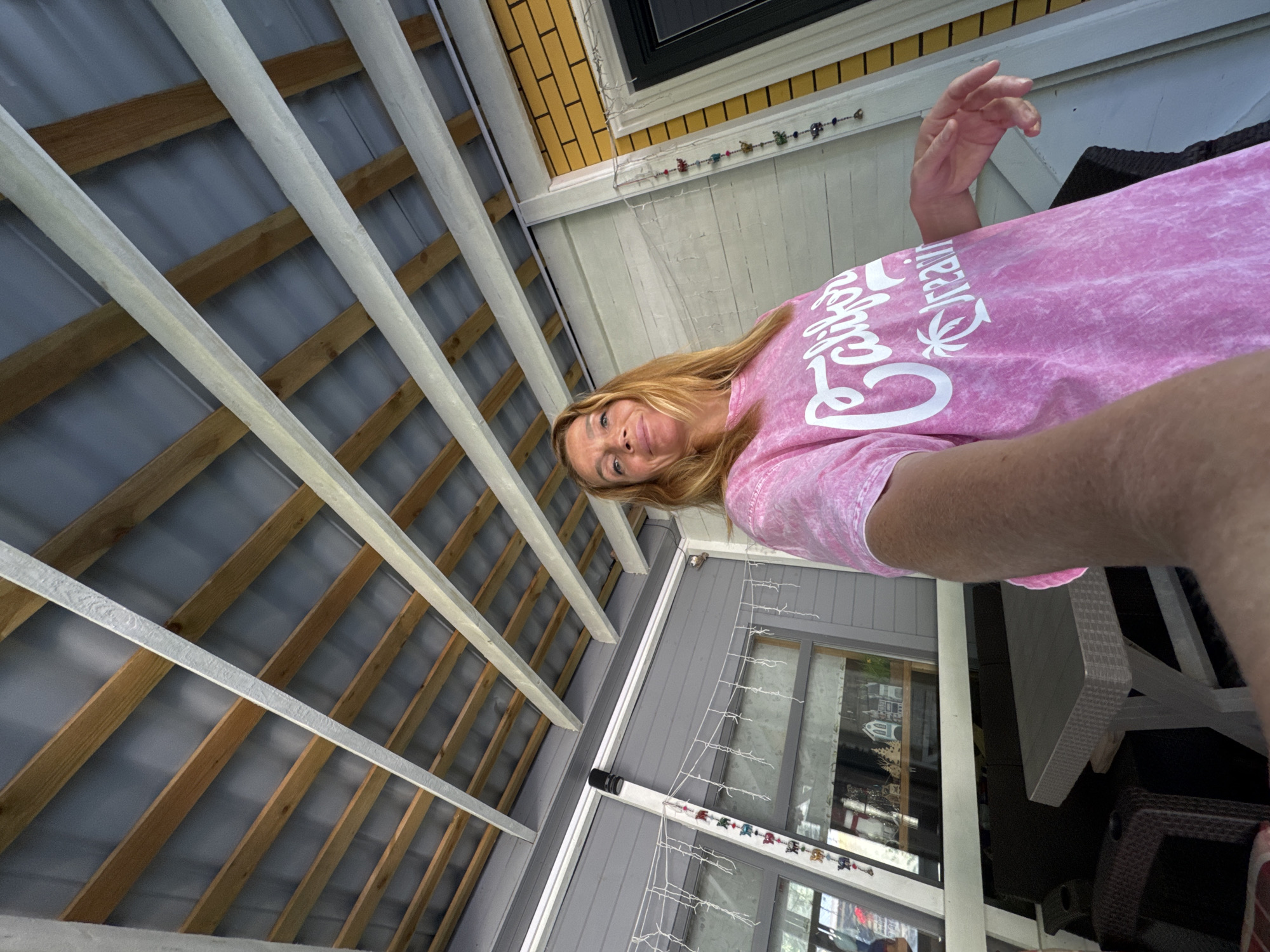 A woman with shoulder-length blonde hair is taking a selfie on a porch. She is wearing a pink tie-dye t-shirt with white text that reads "California Dreaming" and a palm tree graphic. The porch has a gray, slatted roof supported by wooden beams, and a gray siding wall with windows. To the right of the woman is a yellow brick wall with a dark door. There is a table and chairs on the porch, and string lights with decorative charms are hanging from the wall. The woman is smiling slightly and looking at the camera. The scene is outdoors during daylight. The image is taken in Zelenaya Roshcha, Russia.