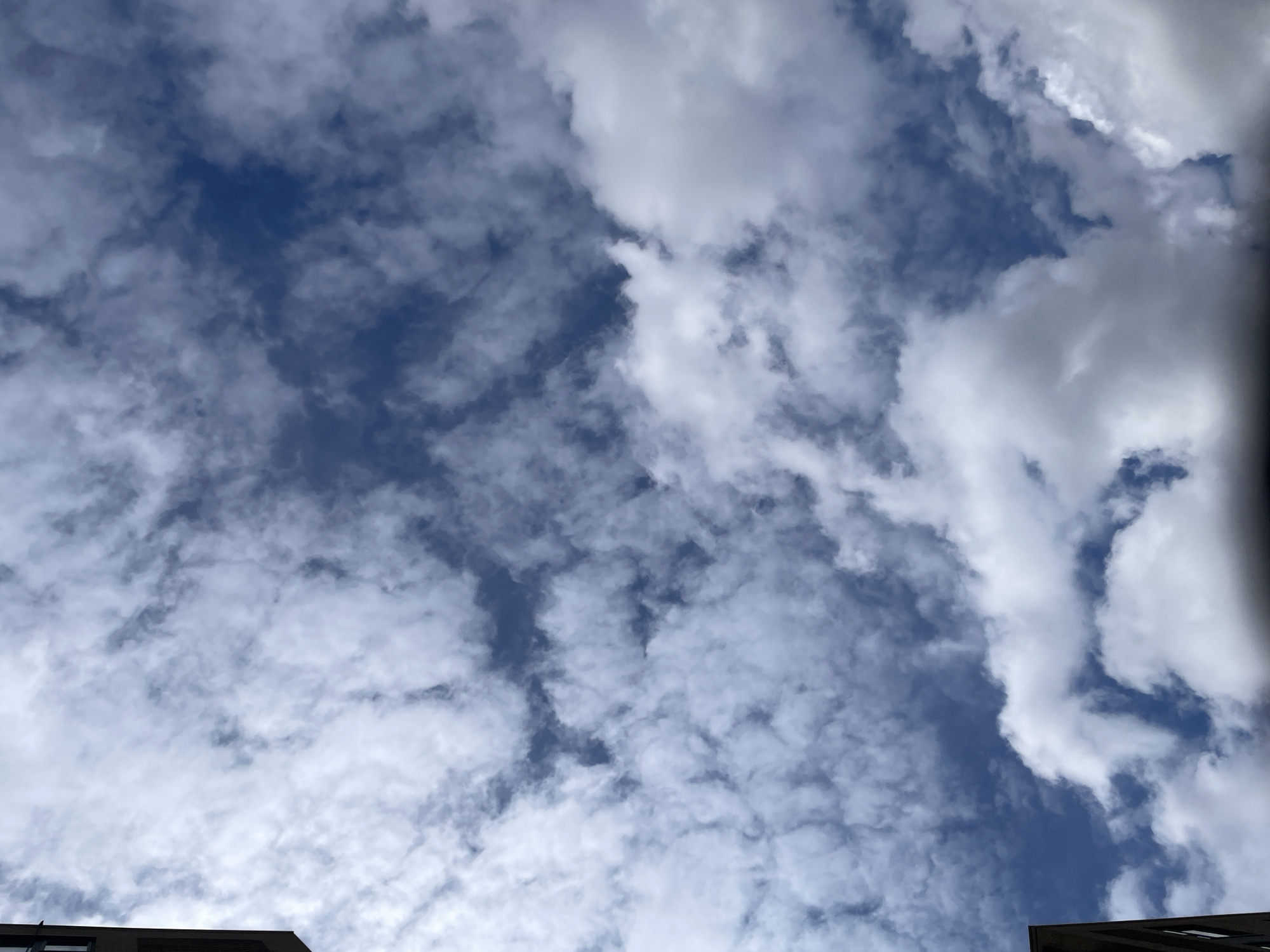 The image is an upward shot towards a cloudy sky. The sky is blue with patches of white clouds scattered throughout. The lower portion of two buildings is visible in the foreground at the bottom of the image. Their appearance suggests a modern, possibly residential or office building. There are no people visible in the image. The image appears to be taken at ground level in an urban environment. It is not possible to conclusively determine the location is in London, United Kingdom based on visual information alone, although architectural elements could be consistent with London's urban landscape.