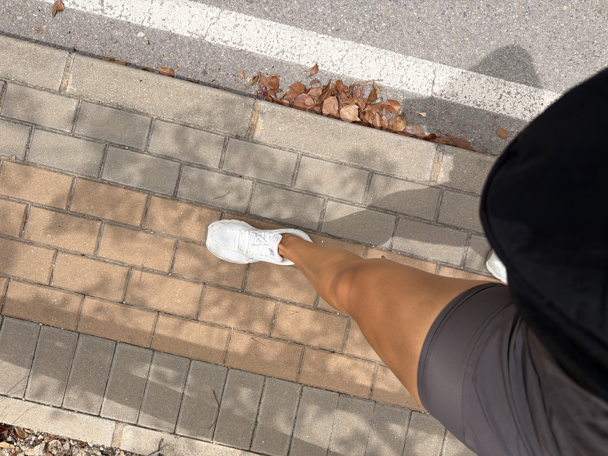 The image shows a person's legs and feet walking on a paved sidewalk. The sidewalk is made of rectangular bricks in alternating shades of grey and tan. To the right of the sidewalk is a road with a white line painted on it. There are some dry leaves on the ground next to the sidewalk and on the road. The person is wearing white sneakers, grey shorts, and a black top or pants. The lighting suggests it is daytime and sunny, with shadows cast by unseen trees. The scene appears to be an outdoor urban or suburban environment.