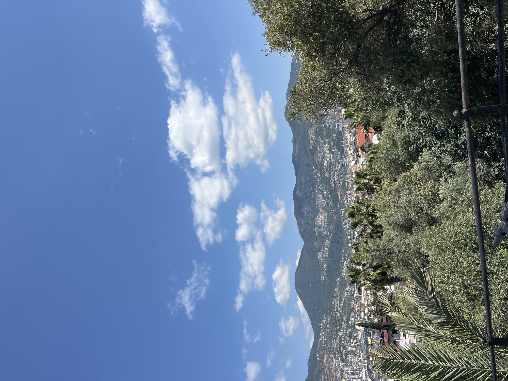 The image shows a view in Alanya, Turkey. In the foreground are green trees and foliage. In the background, there is a mountain range covered with a dense cluster of buildings, indicating a town or city sprawling up the mountainside. The sky is blue and punctuated by puffy white clouds. A fence is visible on the right side of the image.