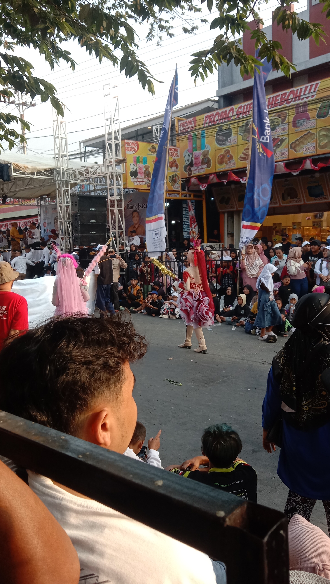 The image depicts an outdoor event in the city of Jatisawit, Indonesia. In the foreground, the backs of several people are visible, suggesting they are spectators. The middle ground is dominated by a street scene with a crowd gathered. At the center of the crowd, a performer in a red and white costume with a ruffled skirt is the main subject, holding a gold-colored staff. To the left, another person in a pink veil and costume is visible. Behind the crowd, there are banners with text, including "PROMO HEBOH!!!" and advertisements for a bank. A stage structure with speakers is set up on the left side of the frame. The background shows buildings and trees, indicating an urban or semi-urban environment. The overall scene appears to be a festive or cultural gathering.