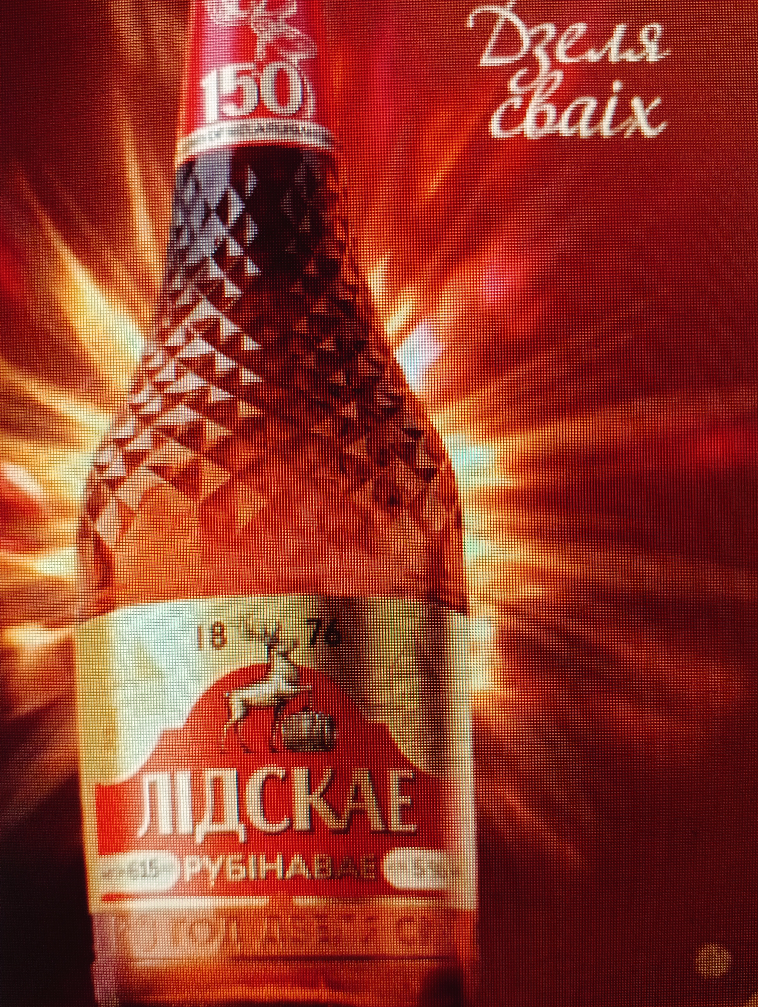 The image is a close-up of a bottle with a red and gold label. The bottle has a textured, faceted design and is illuminated by a strong light source that creates a radiant effect. The label features Cyrillic text and a graphic of a deer. The text "150" is prominent on the neck of the bottle. In the upper right corner, there is more Cyrillic text written in a stylized script.
The image is a promotional advertisement, likely for a beverage, given the bottle and branding. The background is a fiery red with glowing light rays, suggesting warmth or intensity.
While the image itself does not contain specific geographical markers, the text "ЛІДСКАЕ" (Lidskaye) on the label refers to a brand of beer and non-alcoholic beverages produced by the Lidskoye Pivo brewery, located in the city of Lida, Grodno Region, Belarus. The text "РУБІНАВАЕ" (Rubinavae) on the label may refer to a specific product line or flavor. The city of Sluck, Belarus, is not directly represented in the visual elements of this image.