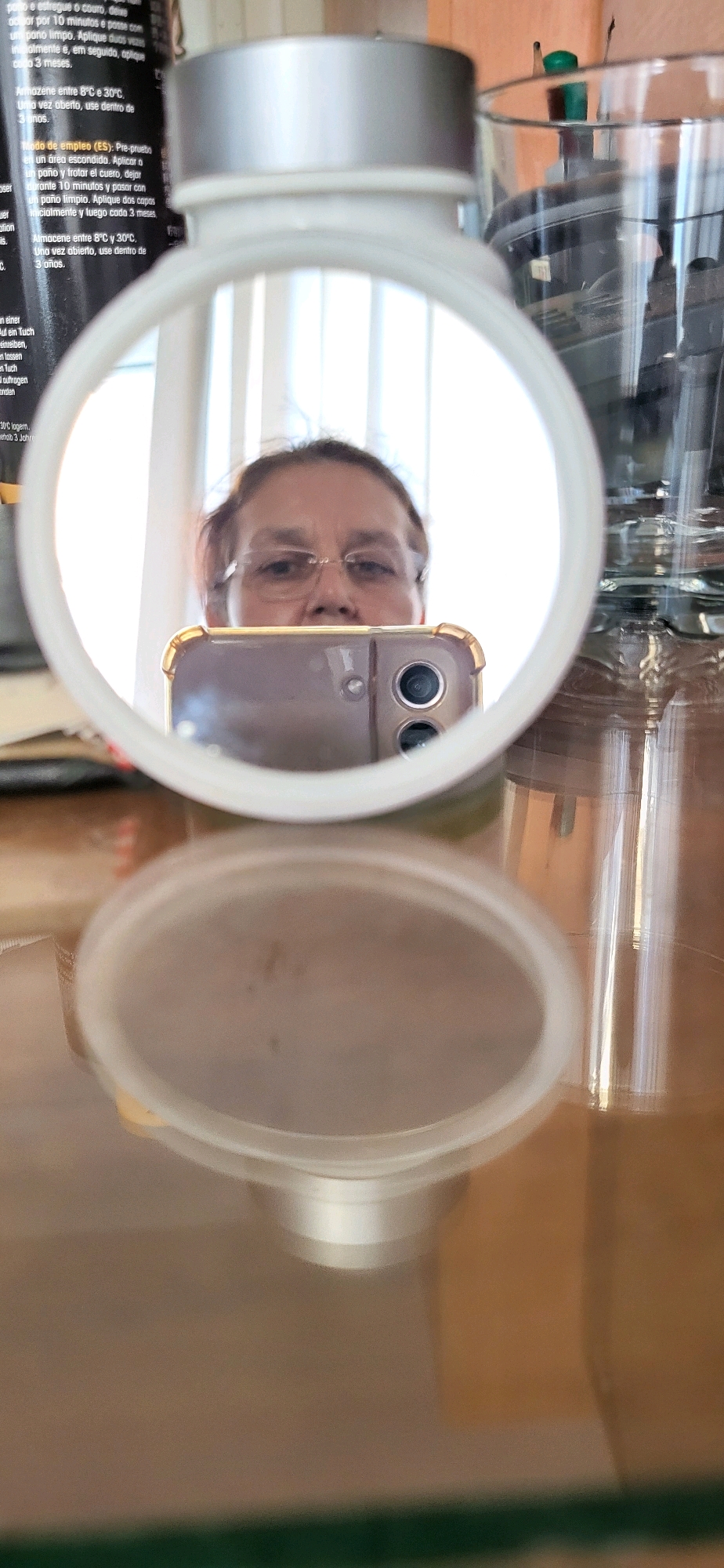 The image shows a reflection in a mirror of a person holding a smartphone to take a selfie. The person has glasses and appears to be looking into the mirror. The smartphone is a light purple color with dual cameras. The reflection also shows part of the person's upper body and the environment around them. In the background, there are glass containers, a black bottle with text, and a bright light source. The scene is set on a table with a reflective surface.
