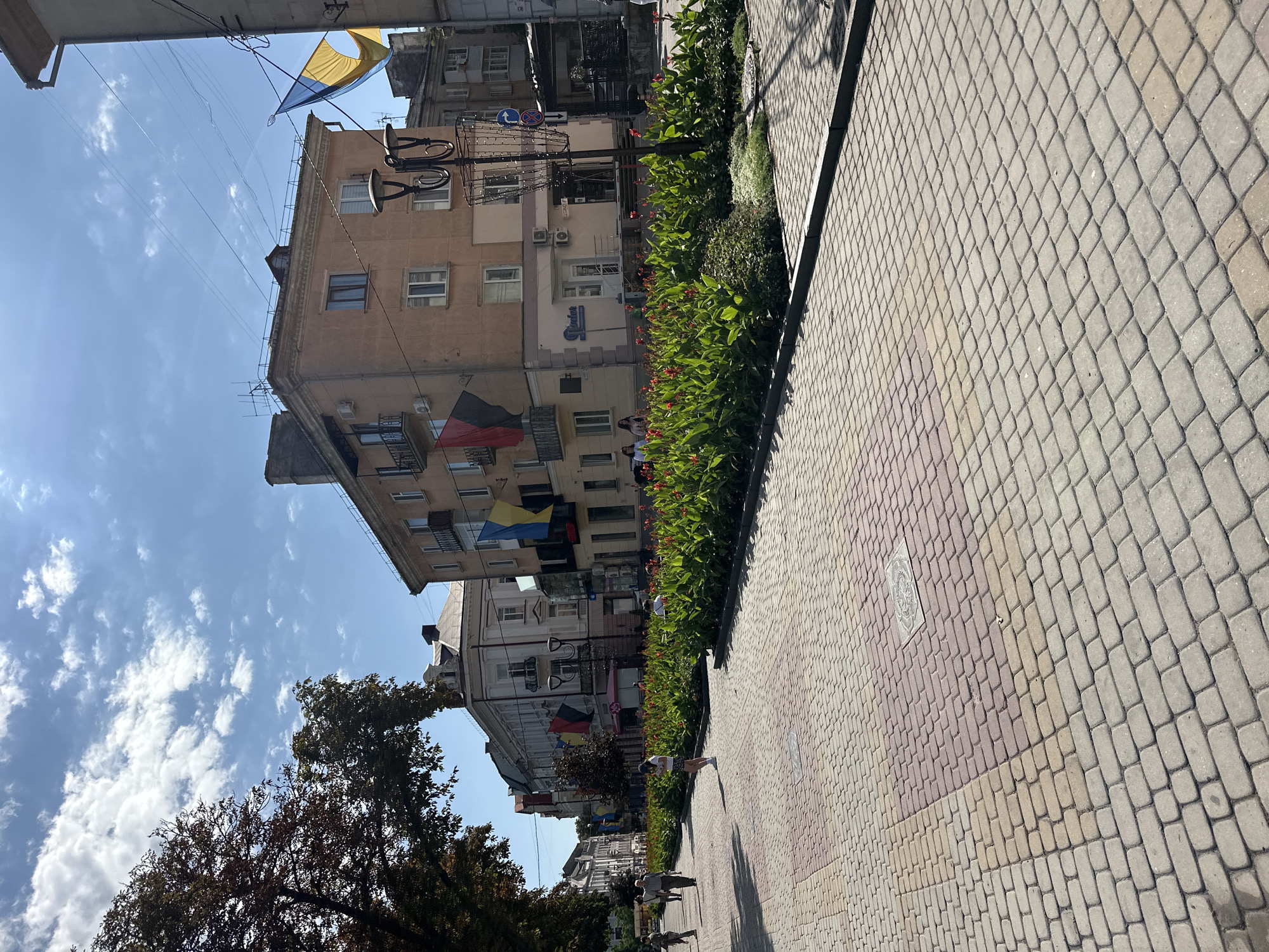 The image depicts a city street in Ternopil, Ukraine, on a sunny day. The street is paved with interlocking pavers, arranged in a pattern of gray, pink, and yellow tones. A central median is filled with lush green plants and vibrant red flowers. Buildings line the street on the right, featuring architectural styles common in Eastern Europe, with balconies and various window designs. Several Ukrainian flags, in blue and yellow, are displayed from the buildings, suggesting a patriotic or celebratory atmosphere. People are seen walking along the sidewalk, some casually strolling, others appearing to be tourists. The sky is blue with scattered white clouds, and the sun casts shadows across the scene. The overall impression is of a lively urban environment on a pleasant day.