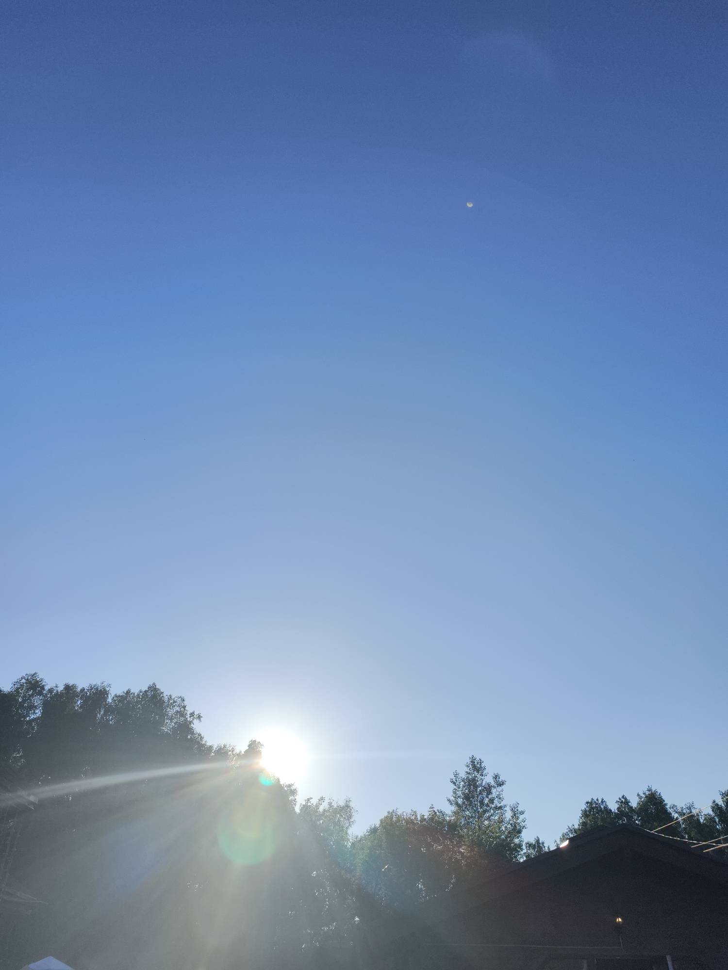 The image shows a bright, clear blue sky. In the upper right quadrant of the sky, a small, crescent moon is visible. The sun is low on the horizon, behind a dense line of trees that span the lower part of the image. The sun is partially obscured by the trees, creating a strong lens flare that radiates outwards. In the bottom right corner of the image, the dark roof of a building is visible, with a small light fixture on its exterior. The overall scene appears to be an outdoor environment, possibly a suburban or rural setting, at either sunrise or sunset.