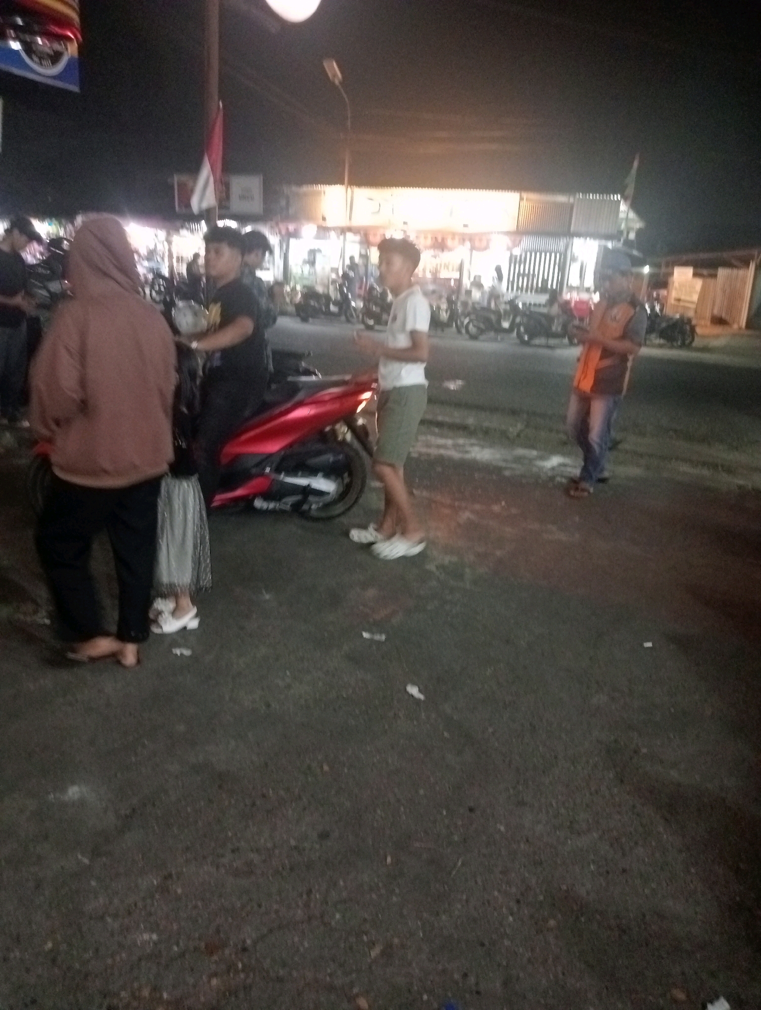 In an outdoor urban setting at night, several individuals are gathered around a red motorcycle parked on an asphalt surface. The scene appears to be near a row of shops with illuminated signs, suggesting a commercial or entertainment district.
A person wearing a brown hooded sweatshirt and black pants stands to the left, facing away from the camera. Beside them, a person in a grey and white patterned skirt and white heels is visible from the waist down.
To the right of the motorcycle, a young man in a white t-shirt and olive green shorts stands with his hands in his pockets. Further to the right, an individual wearing an orange vest over a grey shirt and blue jeans is holding a smartphone.
Several motorcycles are parked in the background, indicating a common mode of transportation. The ground is littered with small white objects, possibly trash or debris. The overall atmosphere is casual and social, with people appearing to be congregating.