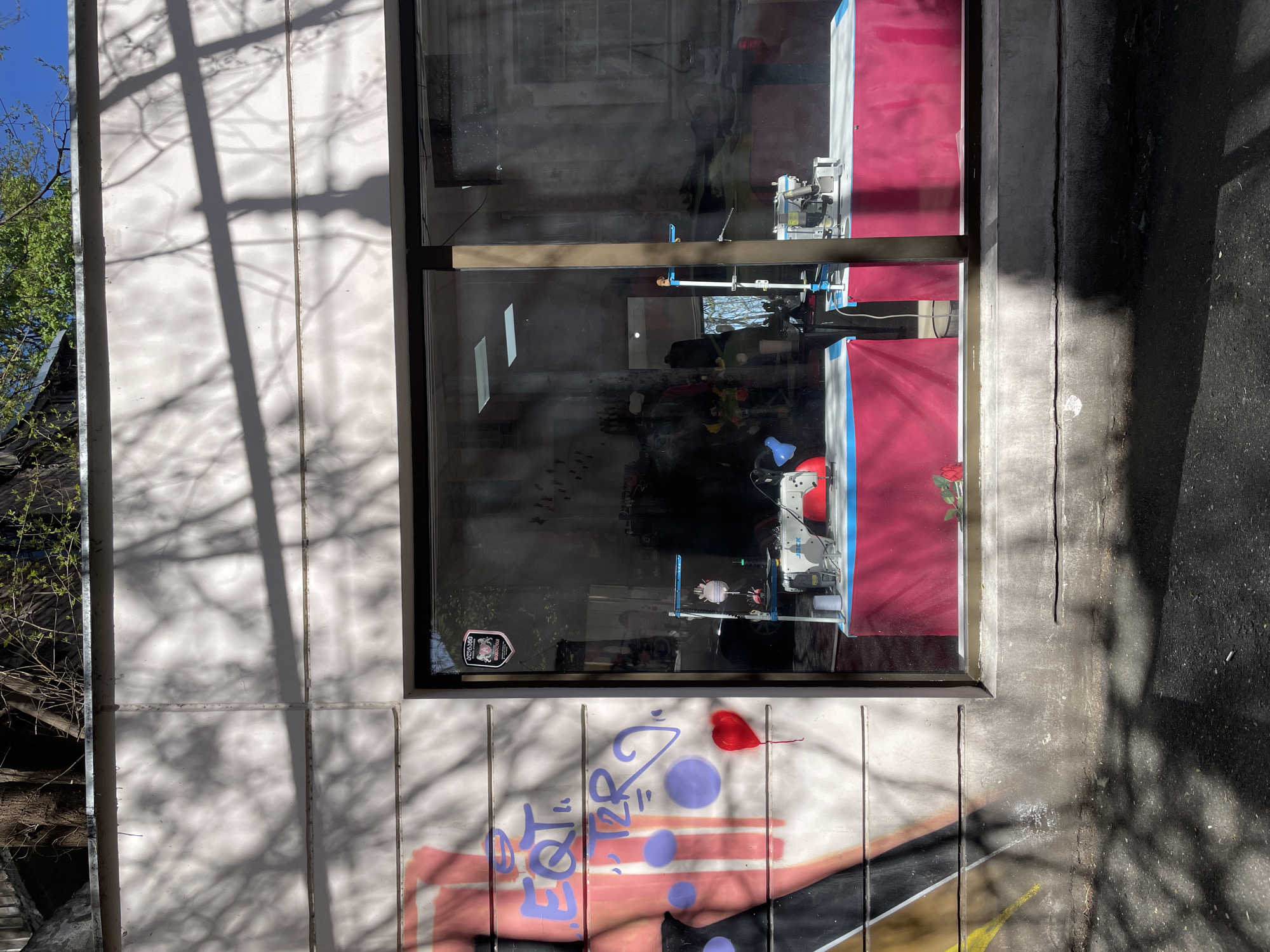A window in a building is seen in Tbilisi, Georgia. The building has graffiti art on the wall underneath the window. Inside the window, there is a sewing machine on a red and white surface. The reflection in the window obscures the objects within the building. The graffiti on the wall is a mix of colors and includes the words "eqt torri". A red heart shape is also painted on the wall. Shadows from trees and branches fall on the wall and the ground beside the building. The background includes some green trees and a blue sky.